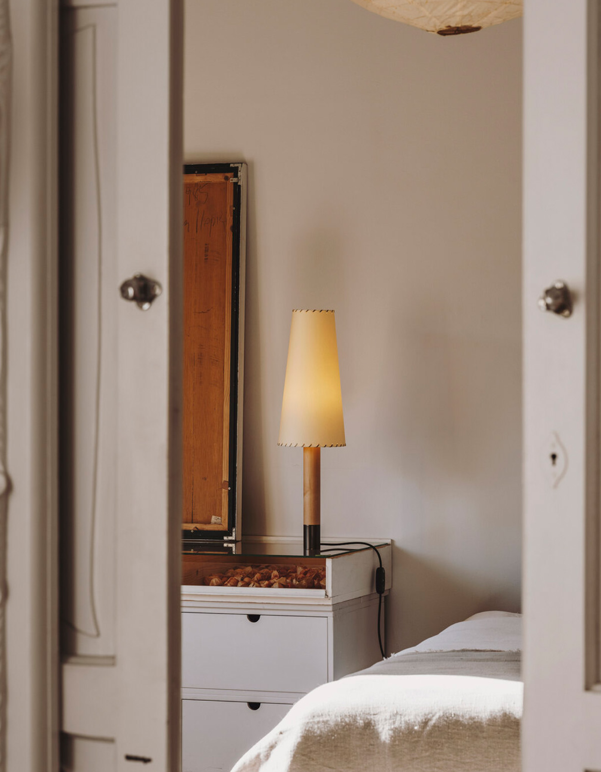 Basica Tall Table Lamp by Santa & Cole with Lighterior. Lightflooded bedroom interior featuring bed, drawer and cylinder shaped table light.