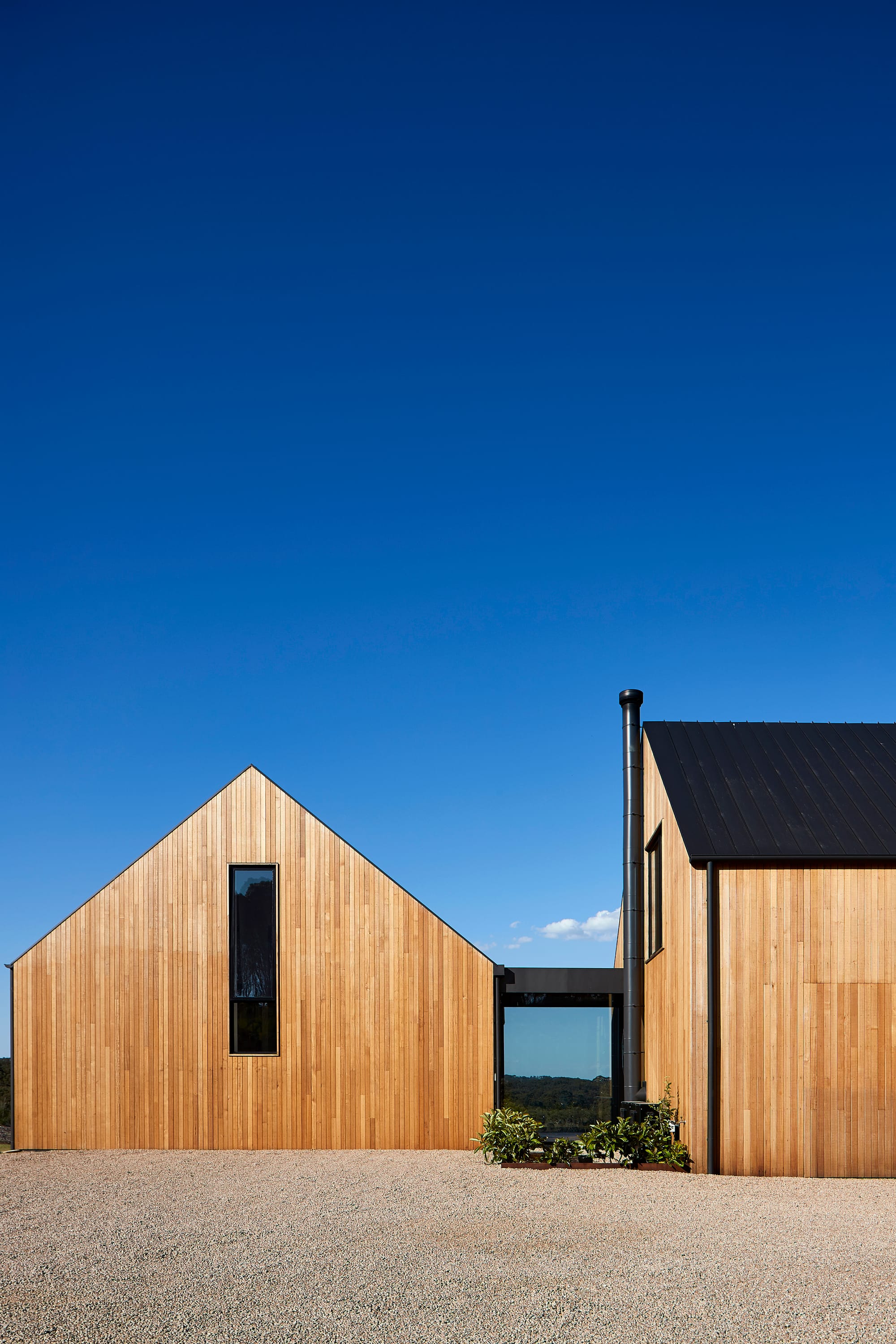 Flinders Residence by Abe McCarthy. Two modern houses featuring gable roof and wood panel facade connected through passerelle with view to surrounding landscape.