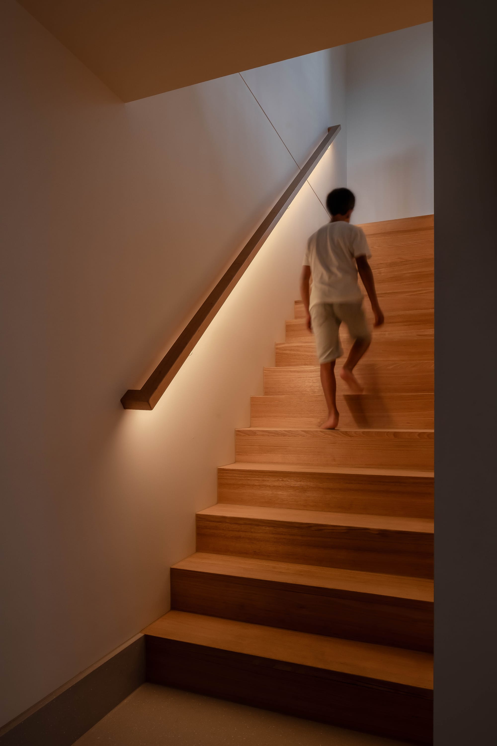 Mahogany House by R Architecture. Wooden staircase featuring illuminated railing opening up into spacious room.