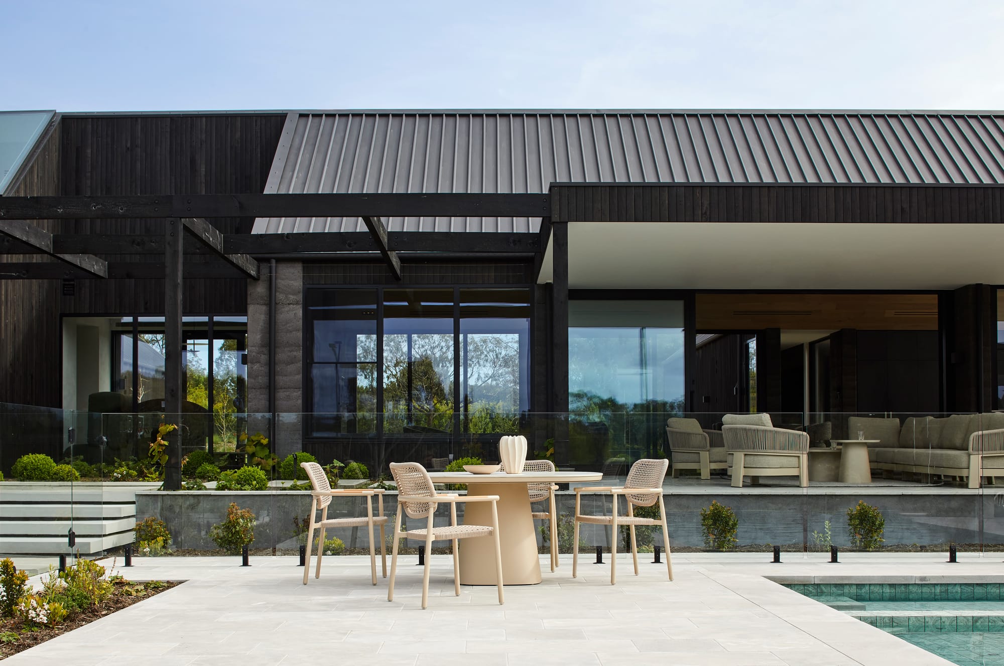 Valencia Curve Dining Table & Valencia Weave Dining Chairs by Globe West. Dining set featuring wooden rattan chairs and table featuring cone shaped base next to pool in front of modern house.