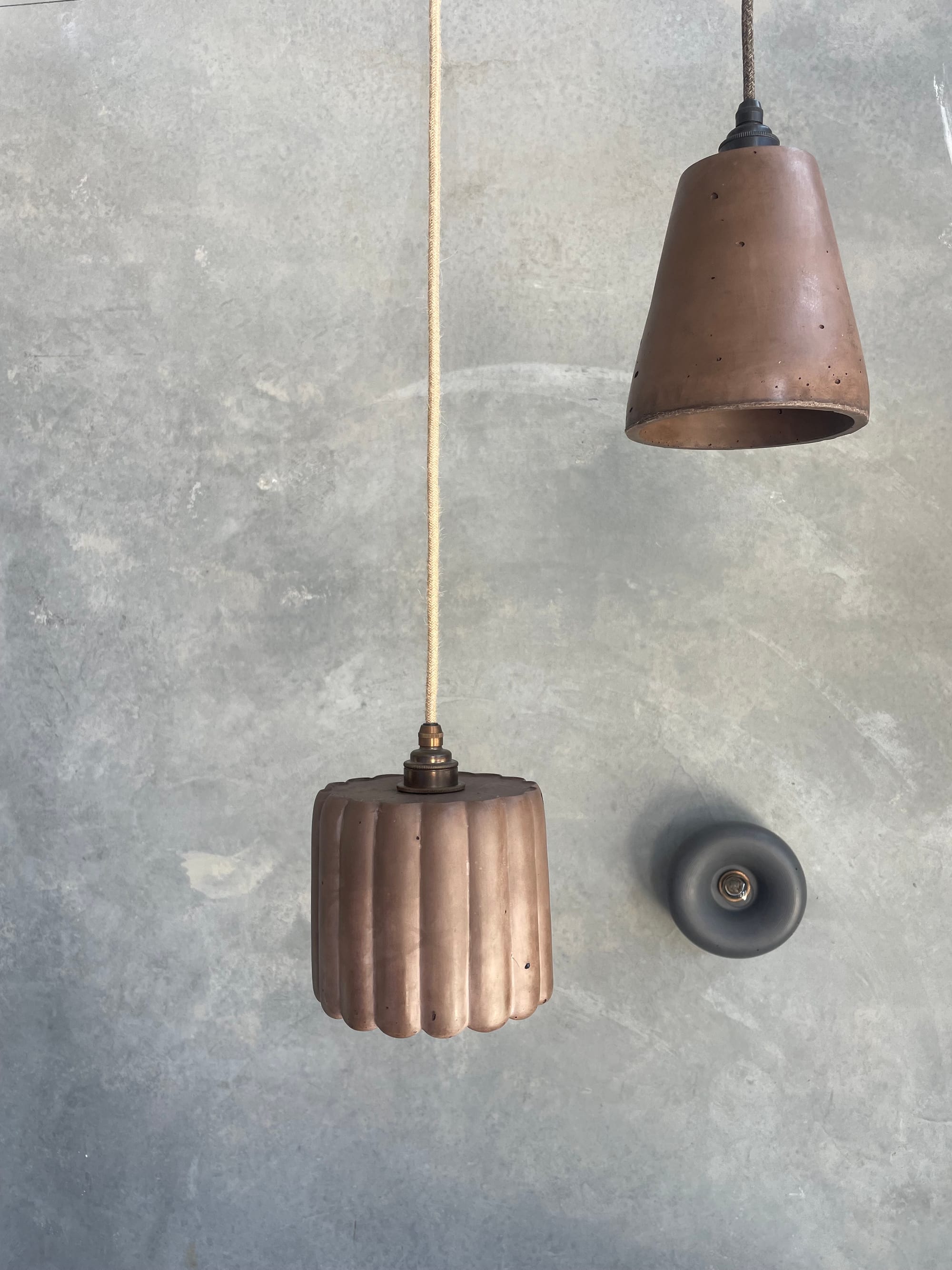 Crete Studio. Left to right: Large Fluted Pendant in Cocoa, Maca Pendant in Cocoa and Haze Wall Sconce in Charcoal.  Two brown concrete pendants hanging on front of concrete wall. Small round grey wall fixture light on right. 