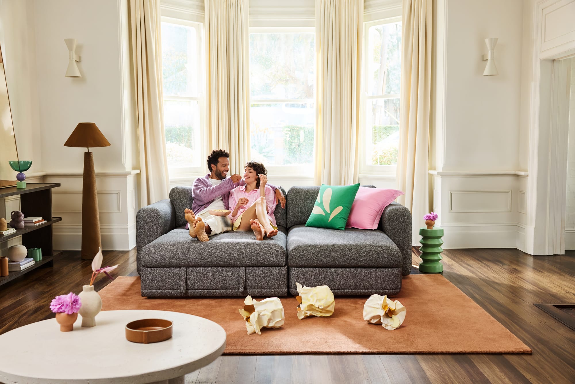 Wanda Sofa Bed by Koala in dark greay showing a couple sitting on the lounge with pink and green cushions