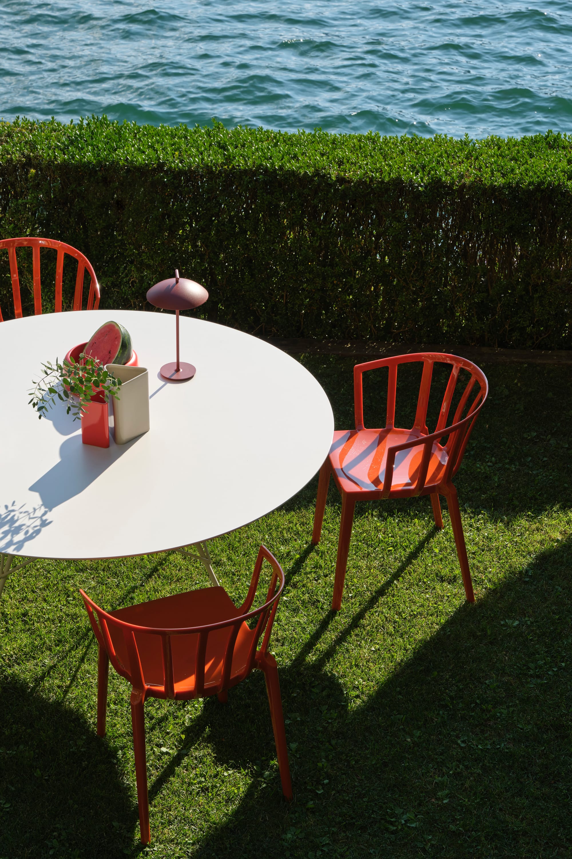 Outdoor Collection Venice Chair by KartellPerth. Red polyurethane outdoor chairs and white round outdoor dining table next to lake and Mediterranean landscape.