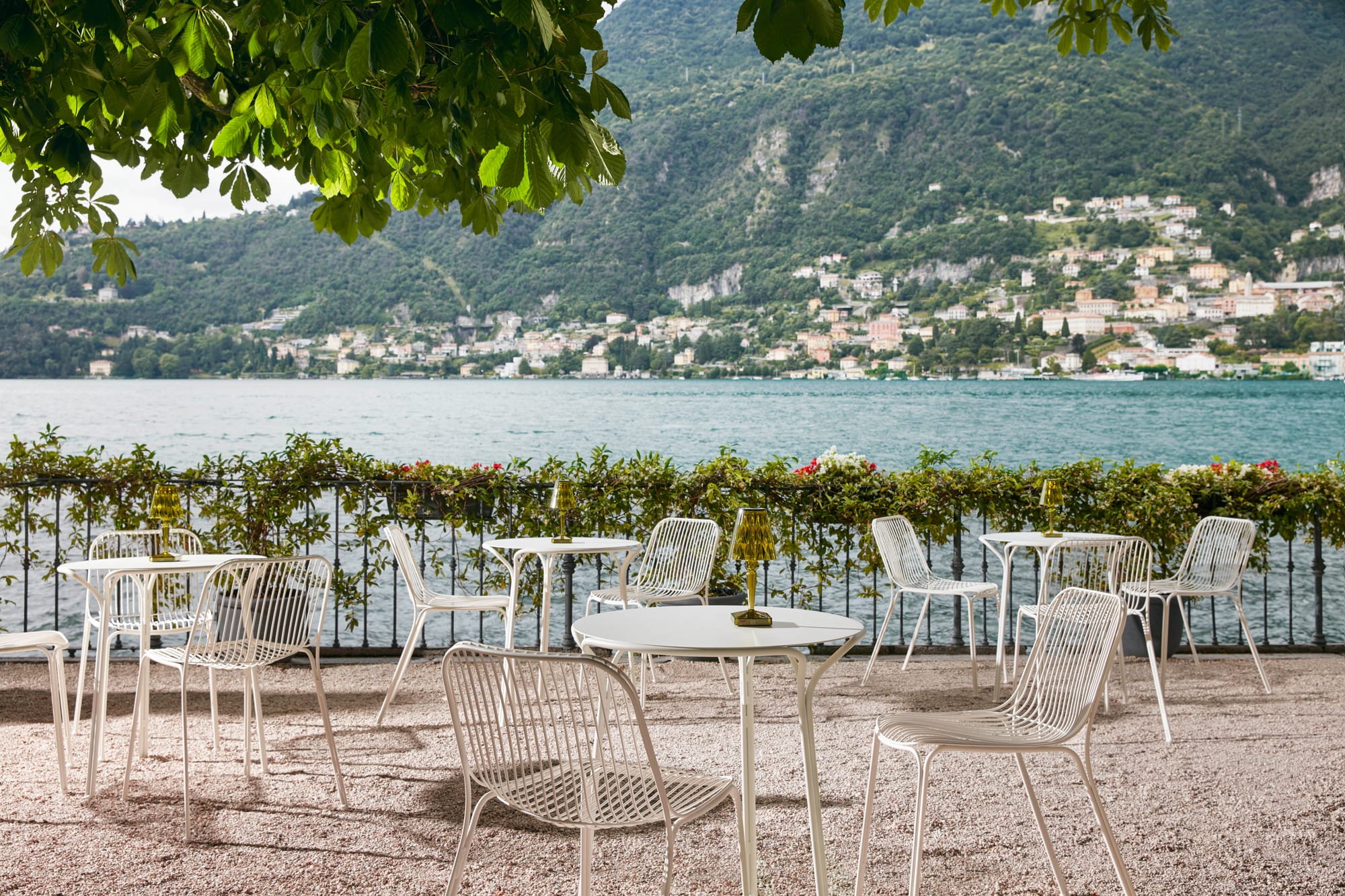 Outdoor Collection Hiray by KartellPerth.Outdoor seating arrangements featuring white metal chairs and table next to Mediterranean lake with view to villages and vegetated mountains.