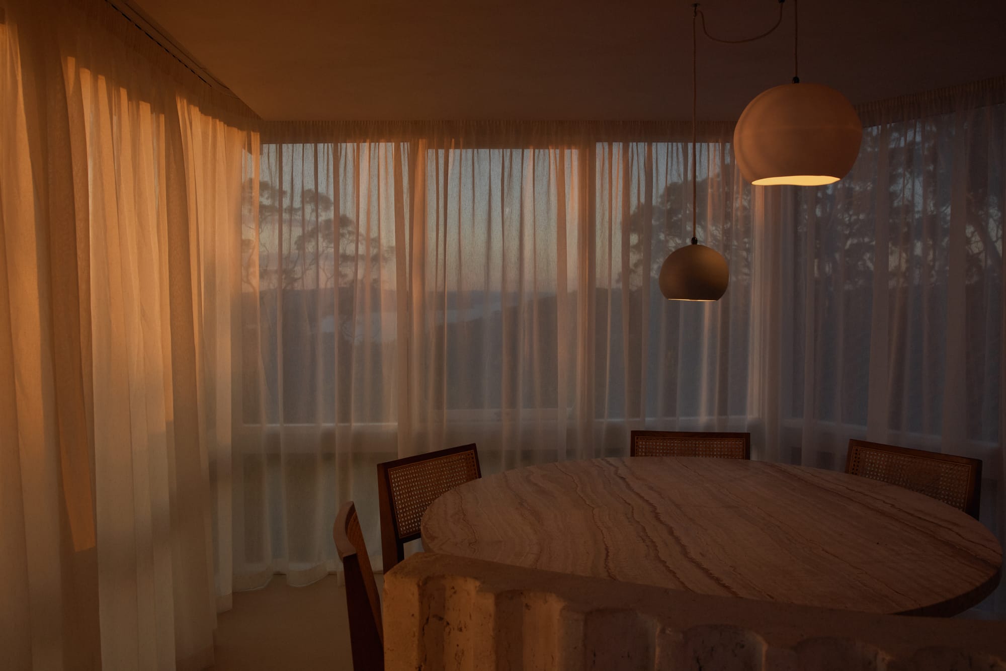 An Orb Pendant Light by Studio Enti hanging over a round, timber dining table with the curtains drawn