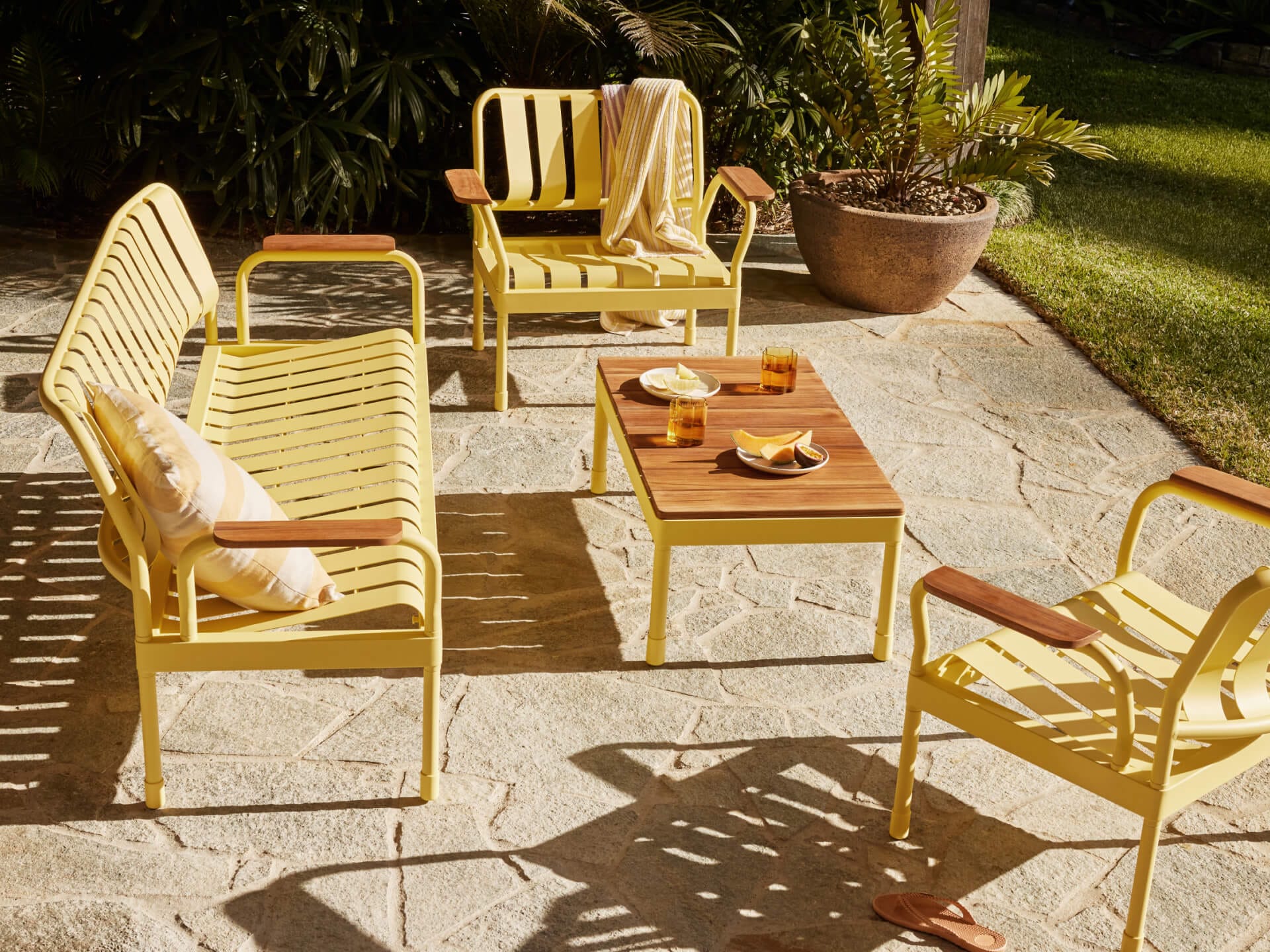 Torakina Outdoor Lounge Set by Koala in Beach Walk. Yellow metal bench seating and dining chairs arranged by timber coffee table on paved surface.