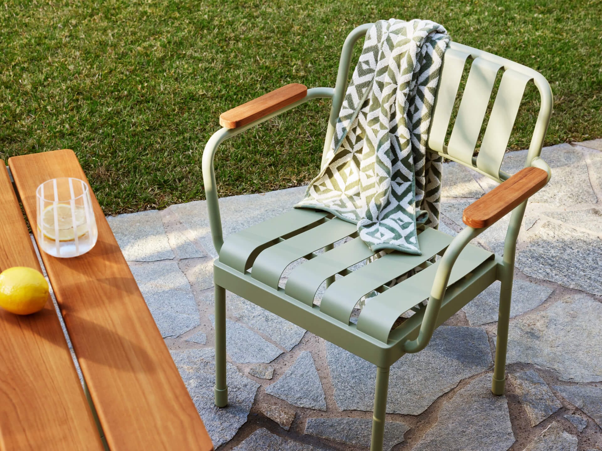 Torakina Outdoor Dining Seating by Koala in Soft Moss. Close up shot of green metal dining chair on paved surface, with grass in background and timber dining table in foreground.