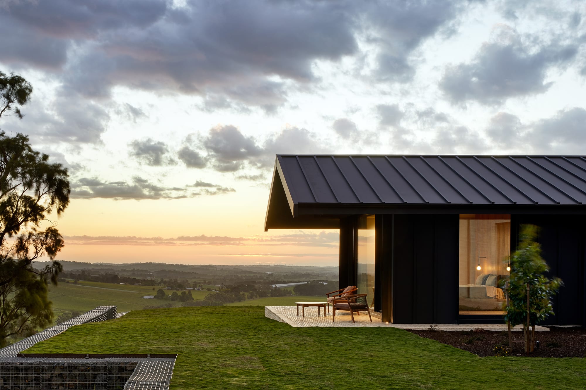 Gruyere Farmhouse by Rachcoff Vella Architecture. Photography by Tatjana Plitt. Exterior of black metal clad home in front of green landscape and sunset. 