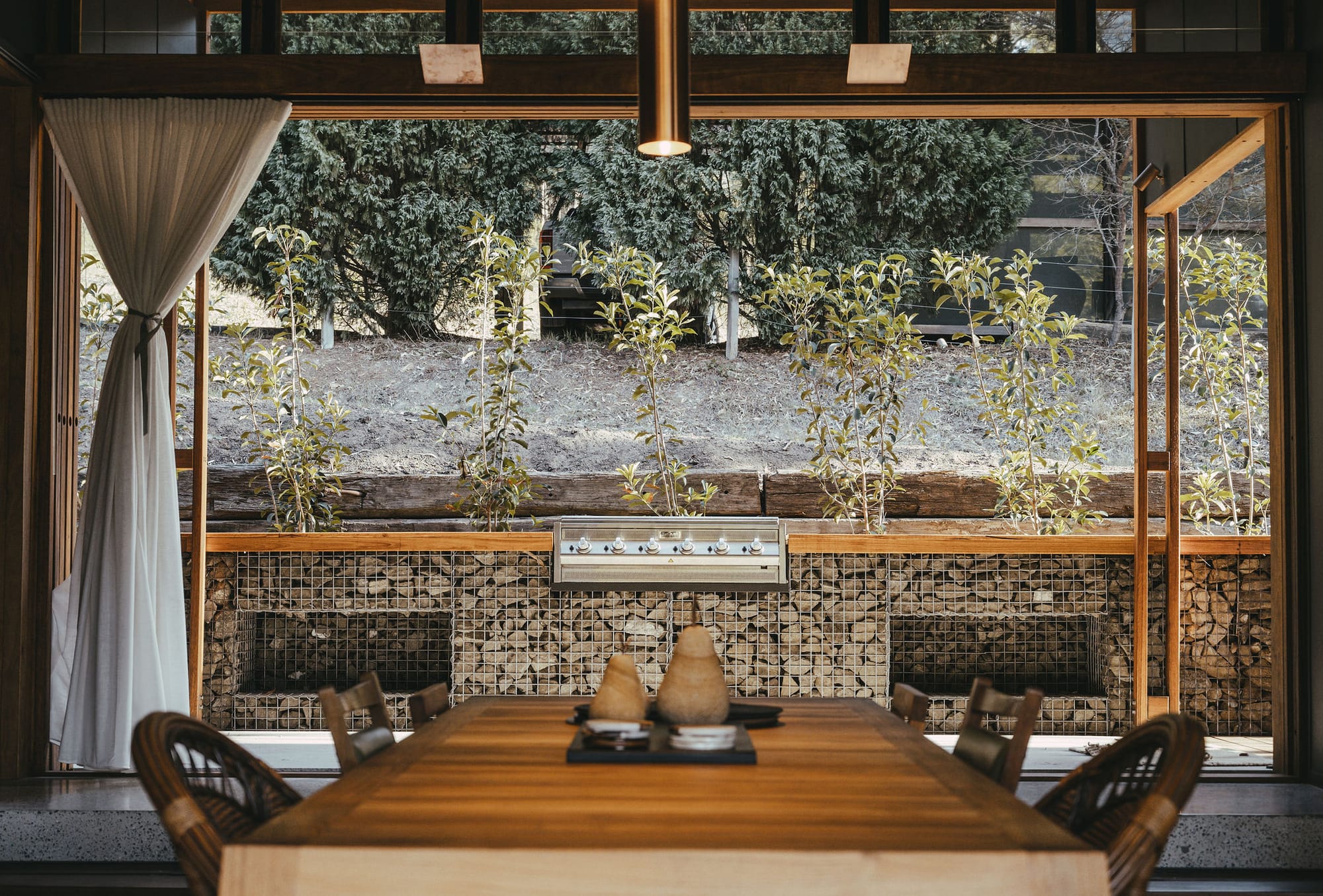 The Summer House at Little Valley Farm. Photography by Milou Hofman Photography. Timber dining table with mismatched dining chairs in foreground with stone bench with integrated barbeque in background.