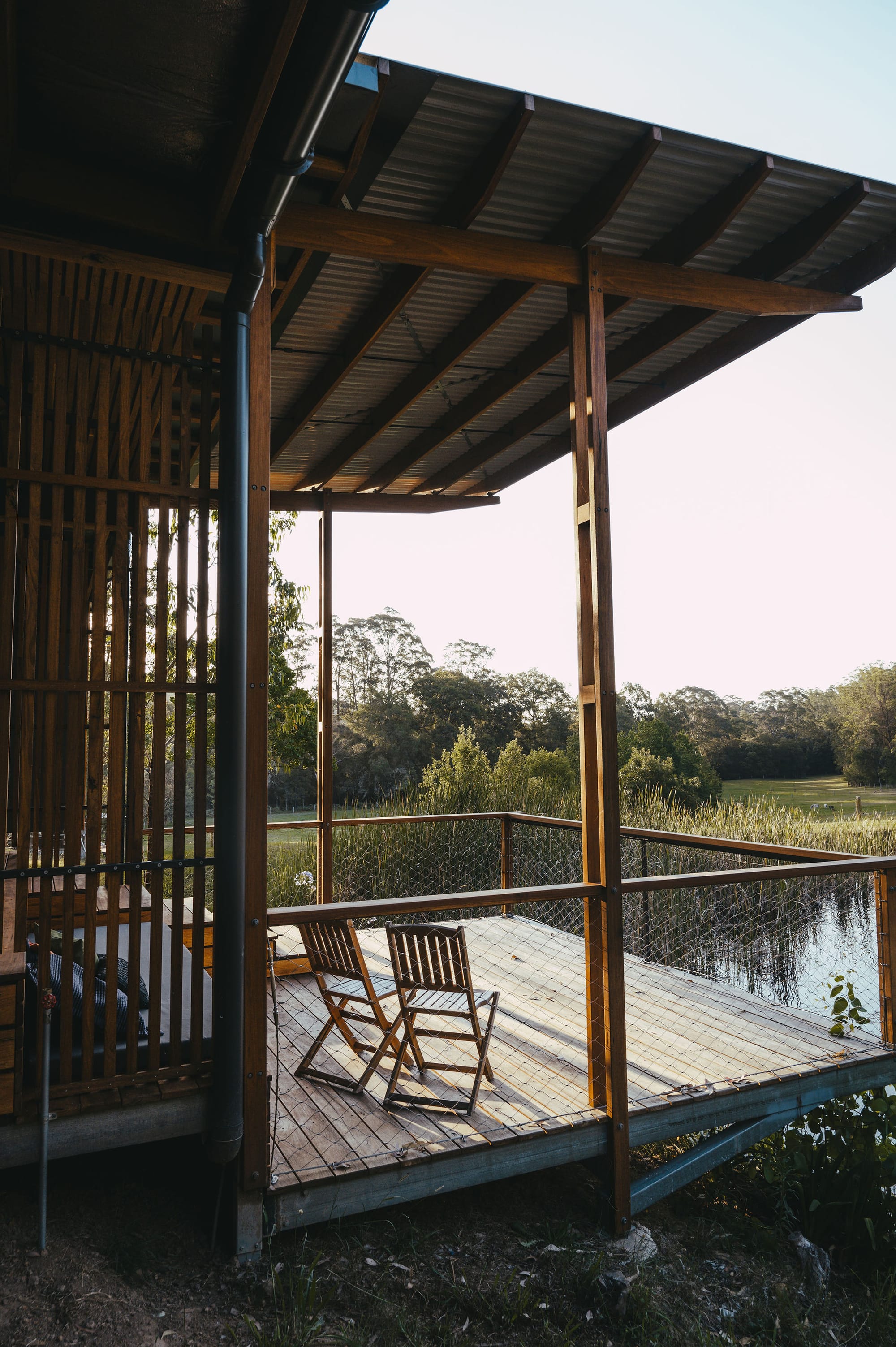 The Summer House at Little Valley Farm. Photography by Milou Hofman Photography. Two timber chairs on timber deck overlooking body of water and large green paddock. 