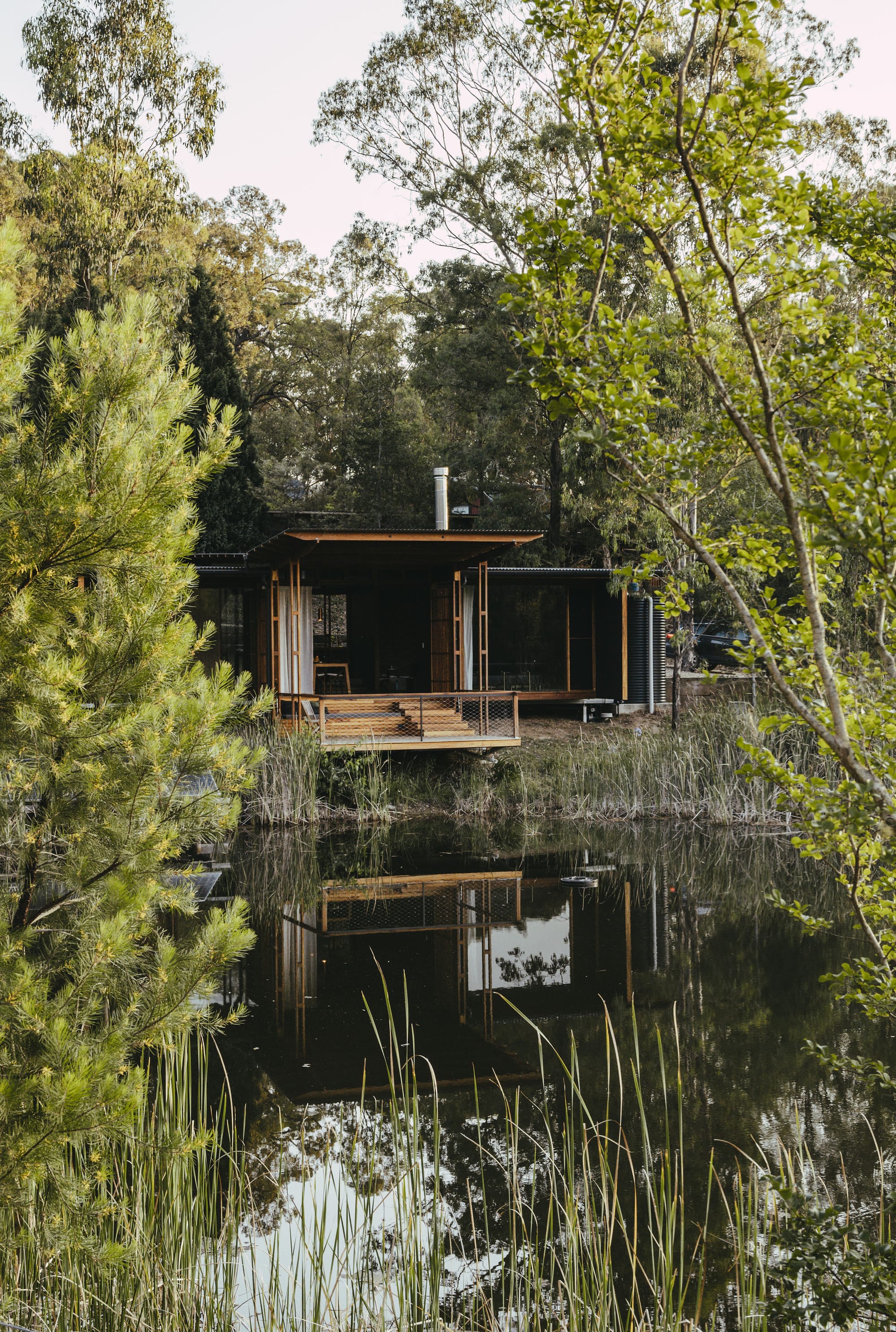 The Summer House at Little Valley Farm. Photography by Milou Hofman Photography. Rear facade of home with timber deck backing onto a large lake, surrounded by native plants. 