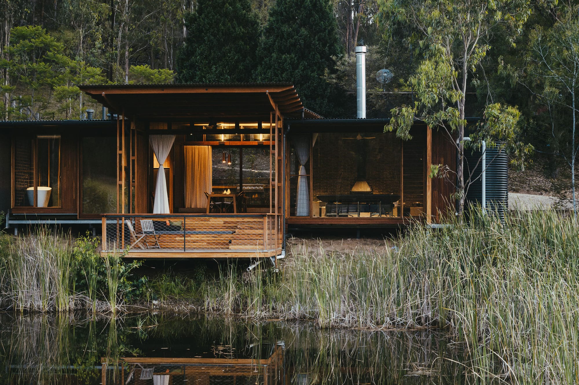 Water fountain in lake surrounded by reed plants, with home with staggered timber deck in background.