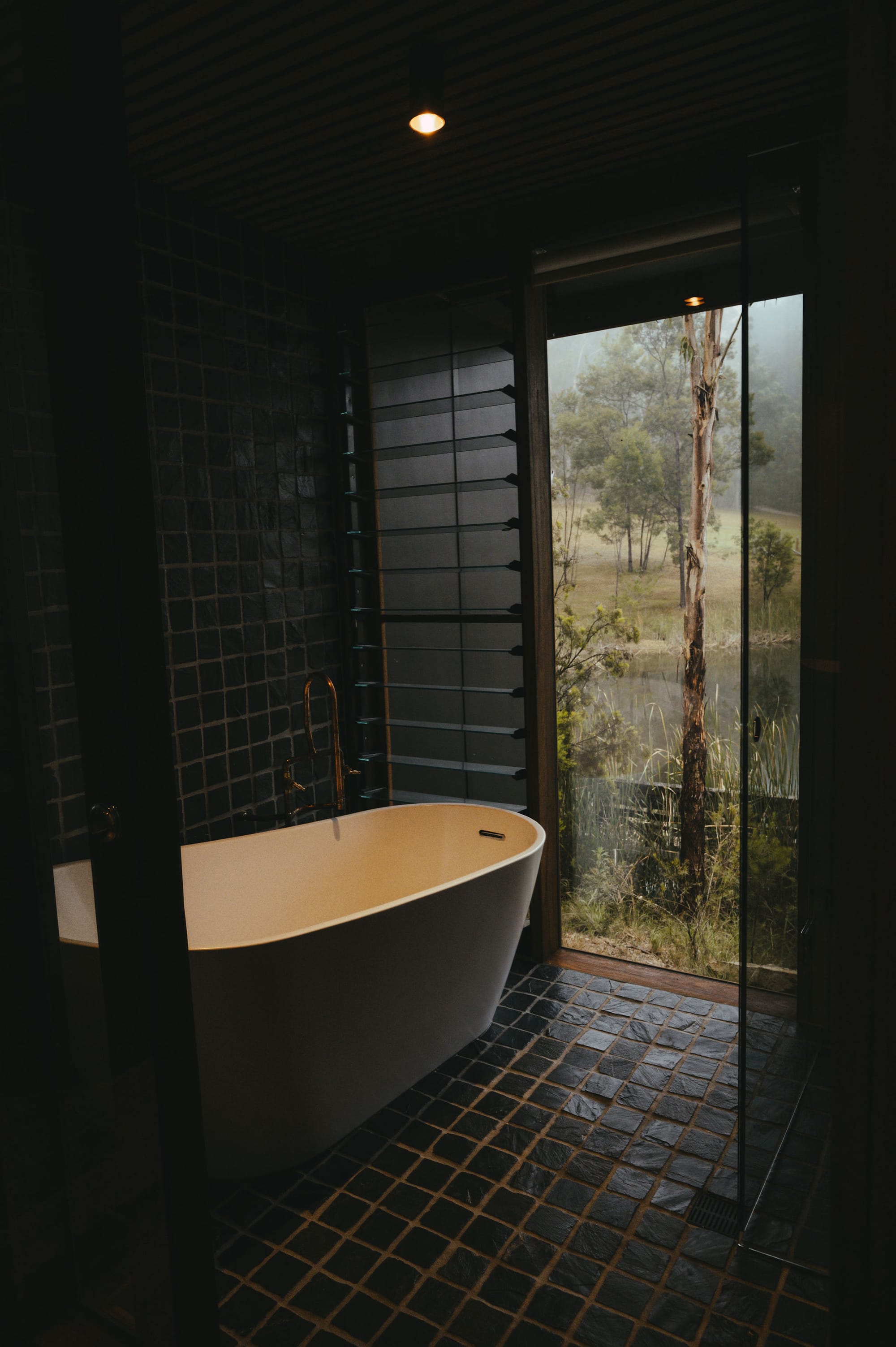 The Summer House at Little Valley Farm. Photography by Milou Hofman Photography. Free standing bath in slate tiled bathroom with full length window overlooking native bush and lake. 