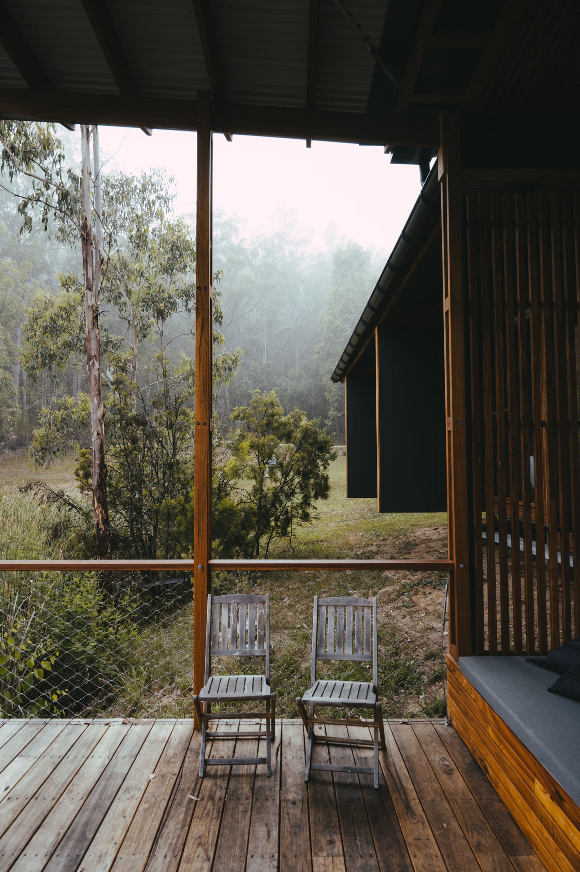 The Summer House at Little Valley Farm. Photography by Milou Hofman Photography. Two worn timber outdoor dining chairs on timber deck, with timber and mesh fencing behind and backdrop of native plantlife and mist.