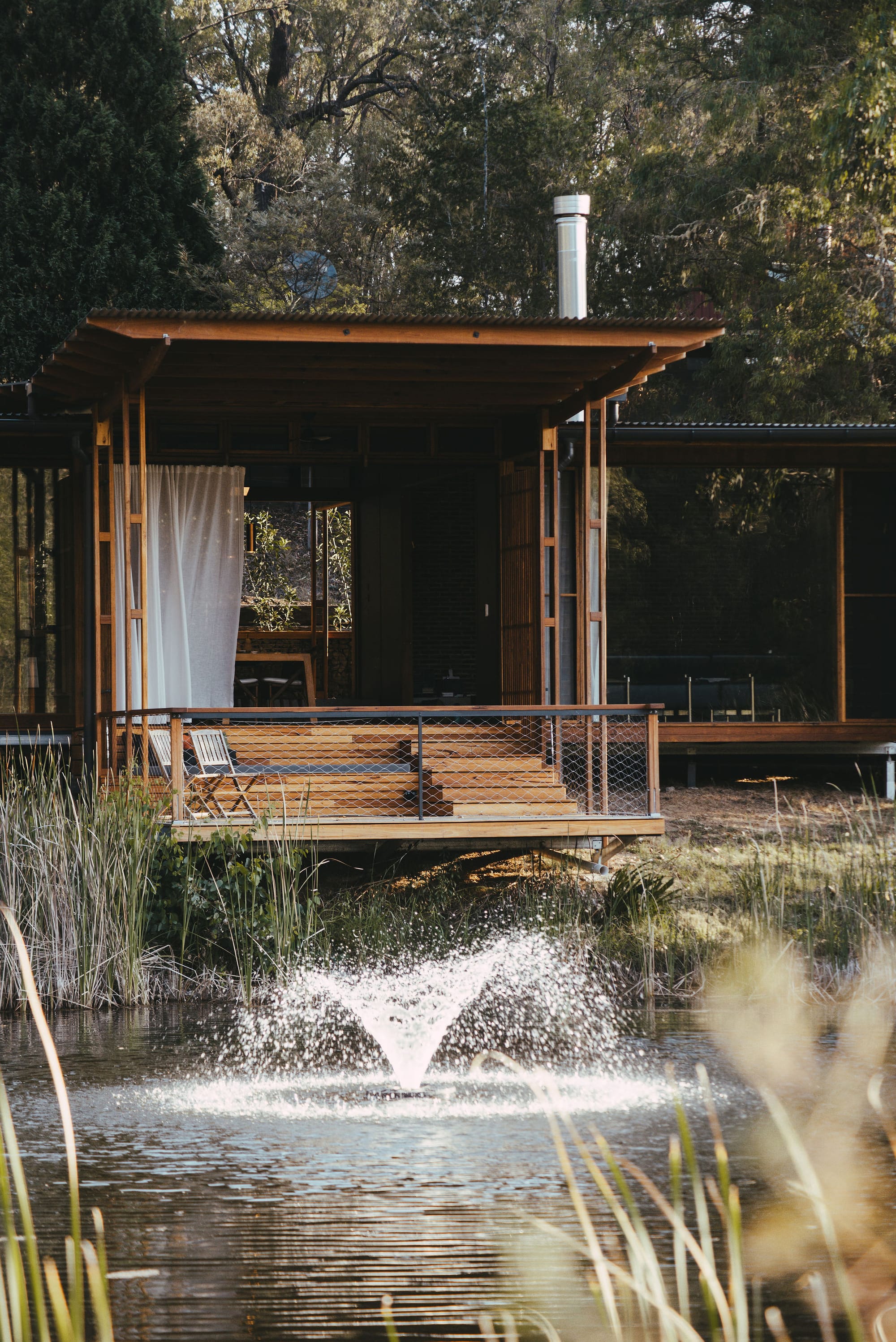 The Summer House at Little Valley Farm. Photography by Milou Hofman Photography. Water fountain in lake surrounded by reed plants, with home with staggered timber deck in background.