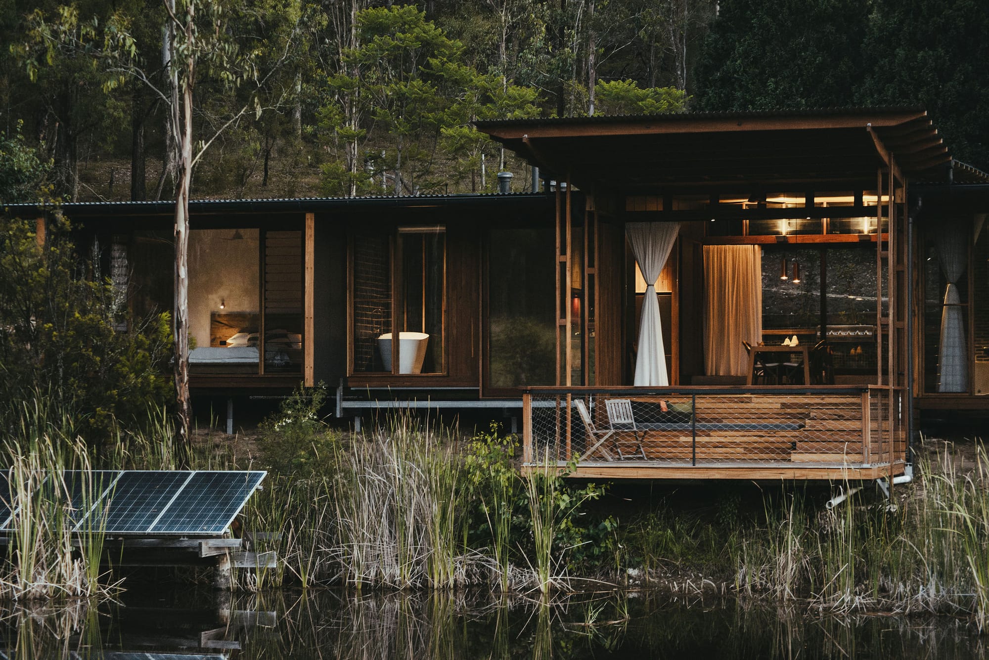 The Summer House at Little Valley Farm. Photography by Milou Hofman Photography. Rear facade of home backing into body of water, constructed from timber with large deck. Surrounded by plants.