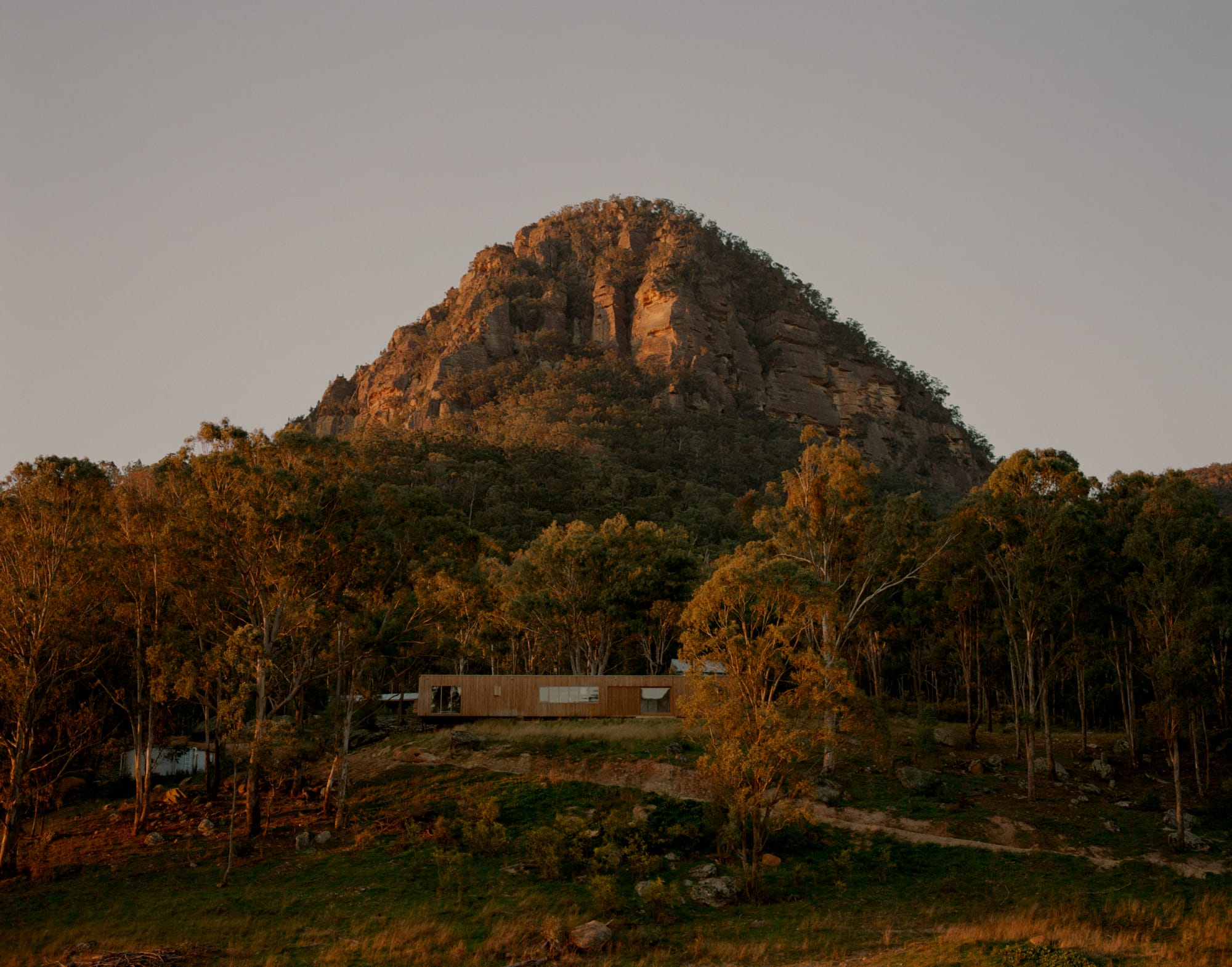 Practice Ground. Photography by Saskia Wilson. Long rectangular residential home clad in timber in foreground of large mountain covered with trees and rocks. 