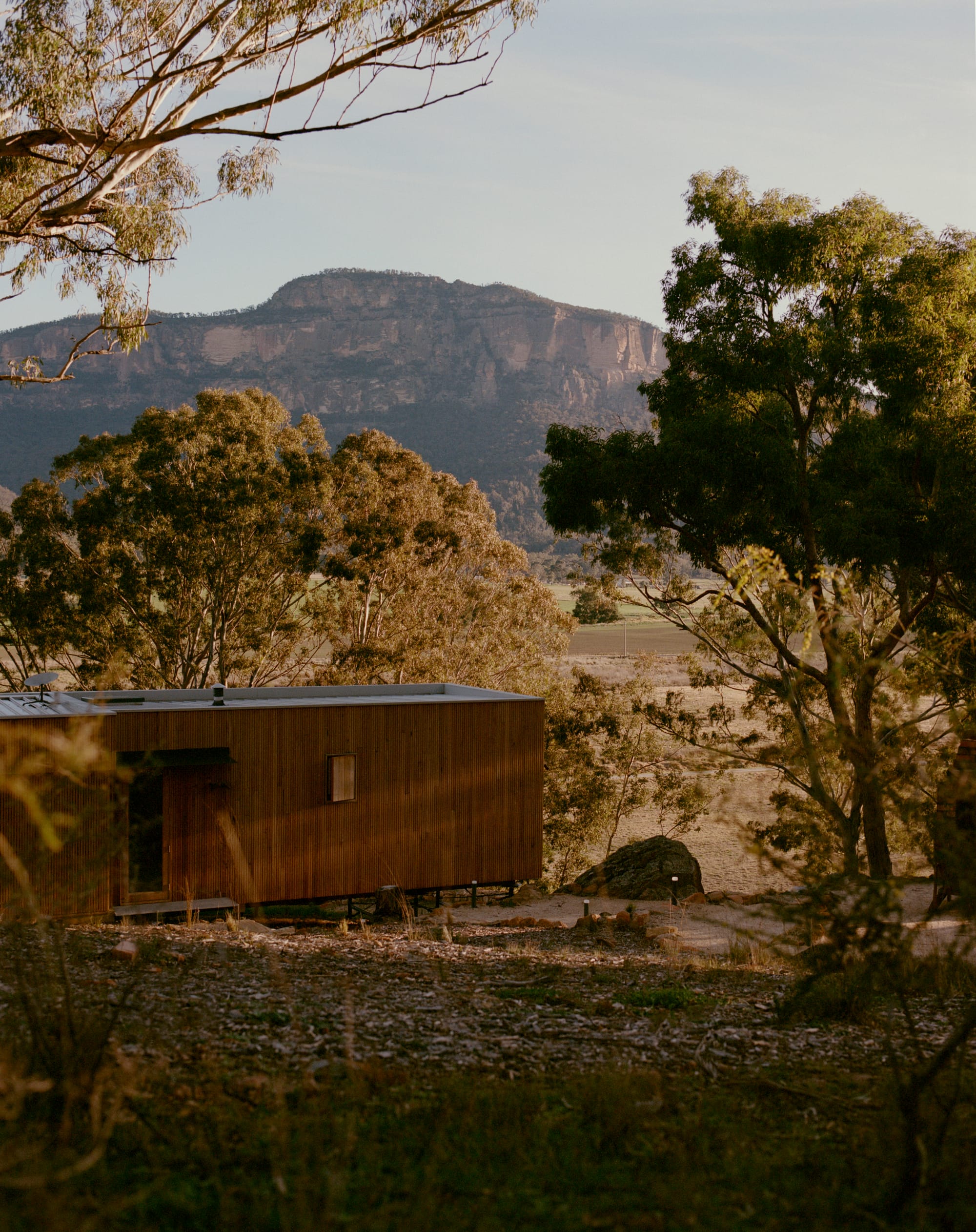 Practice Ground. Photography by Saskia Wilson. Landscape image of tmber clad rectangular modular home in rustic Australian bushland setting with large mountain in the background. 