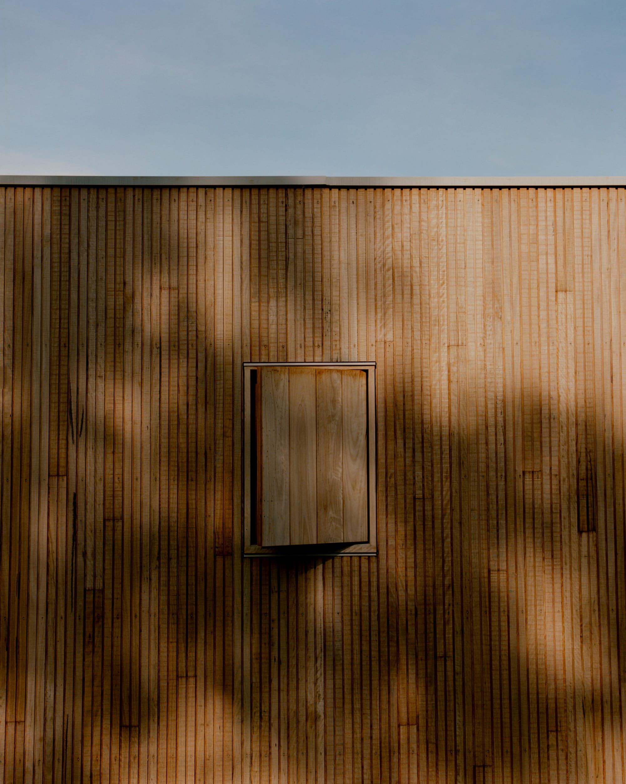 Practice Ground. Photography by Saskia Wilson. Hallway with light tinber clad floors, walls, ceiling, joinery and doors opening onto a bedroom with striped bed linen and small square window. 