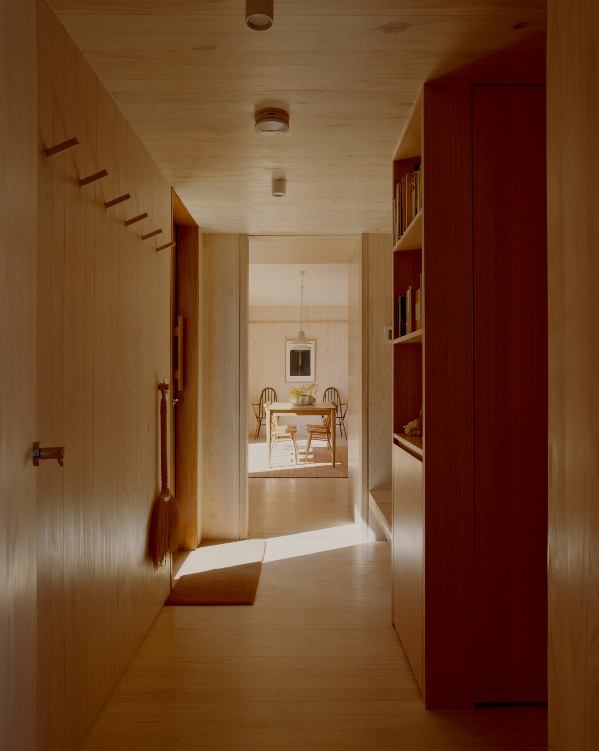 Practice Ground. Photography by Saskia Wilson. Shadowy hallway with timber floors, ceilings, walls, integrated bookshelf and joinery opening onto a timber dining table. 