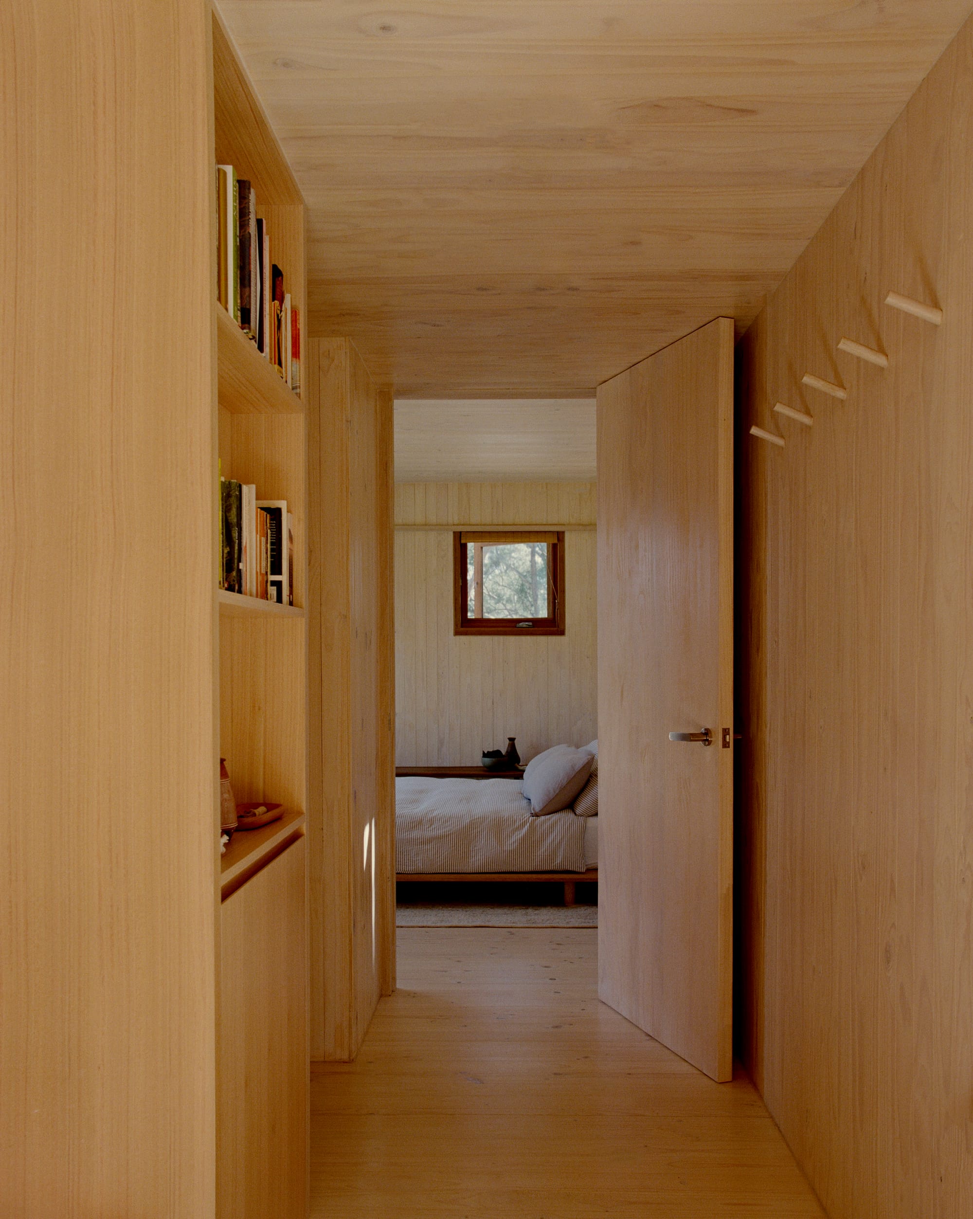 Practice Ground. Photography by Saskia Wilson. Hallway with light tinber clad floors, walls, ceiling, joinery and doors opening onto a bedroom with striped bed linen and small square window. 