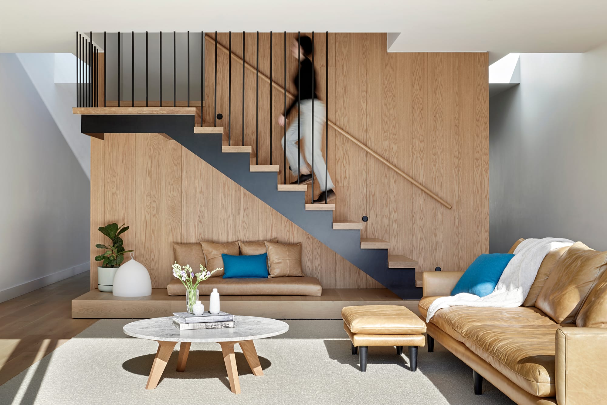 HMAS L02 by Rachcoff Vella Architecture. Photography by Tatjana Plitt. Modern staircase in background of living space with timber walls, light tan couch and blue decor accents. 