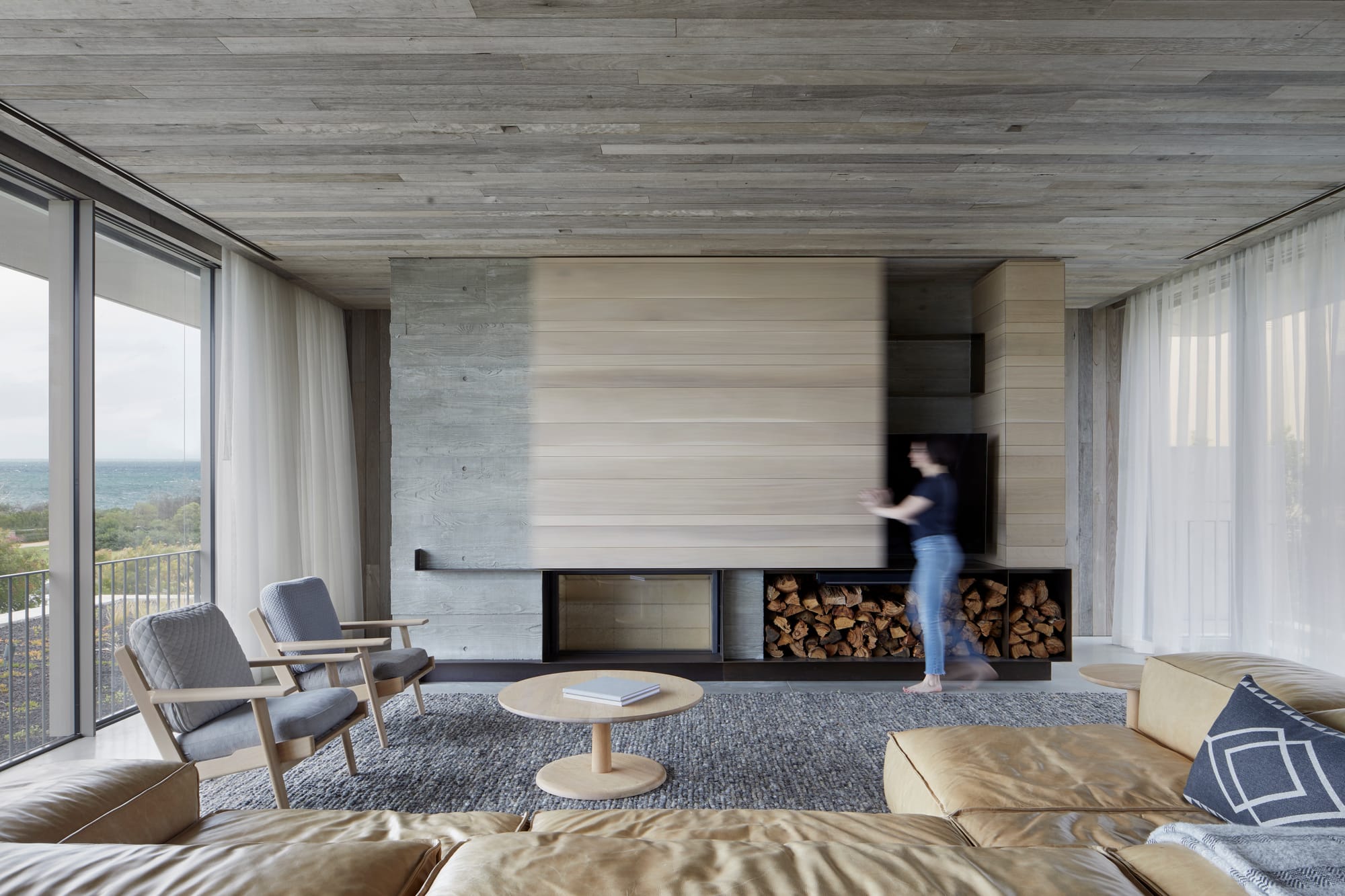 Silver Linings by Rachcoff Vella Architecture. Photography by Tatjana Plitt. Living space with concrete walls and ceiling, tan leather couch and central fireplace with timber cladding. 