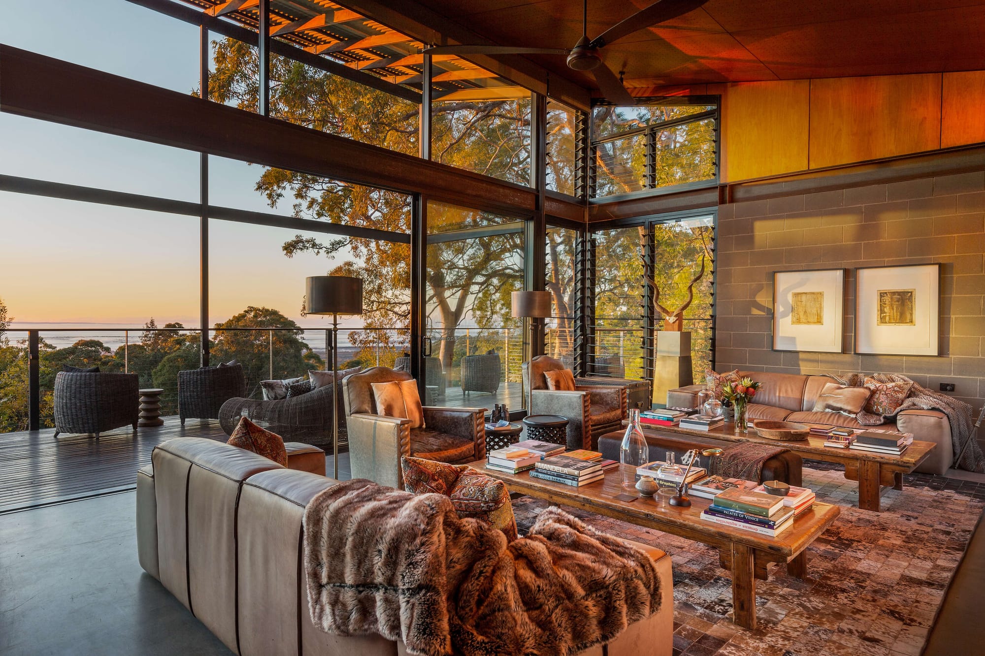 Spicers Sangoma by Barbara Tarnawski Architects. Living space opening onto raised timber deck at sunset, with warm lighting inside cosy and cluttered interior. 