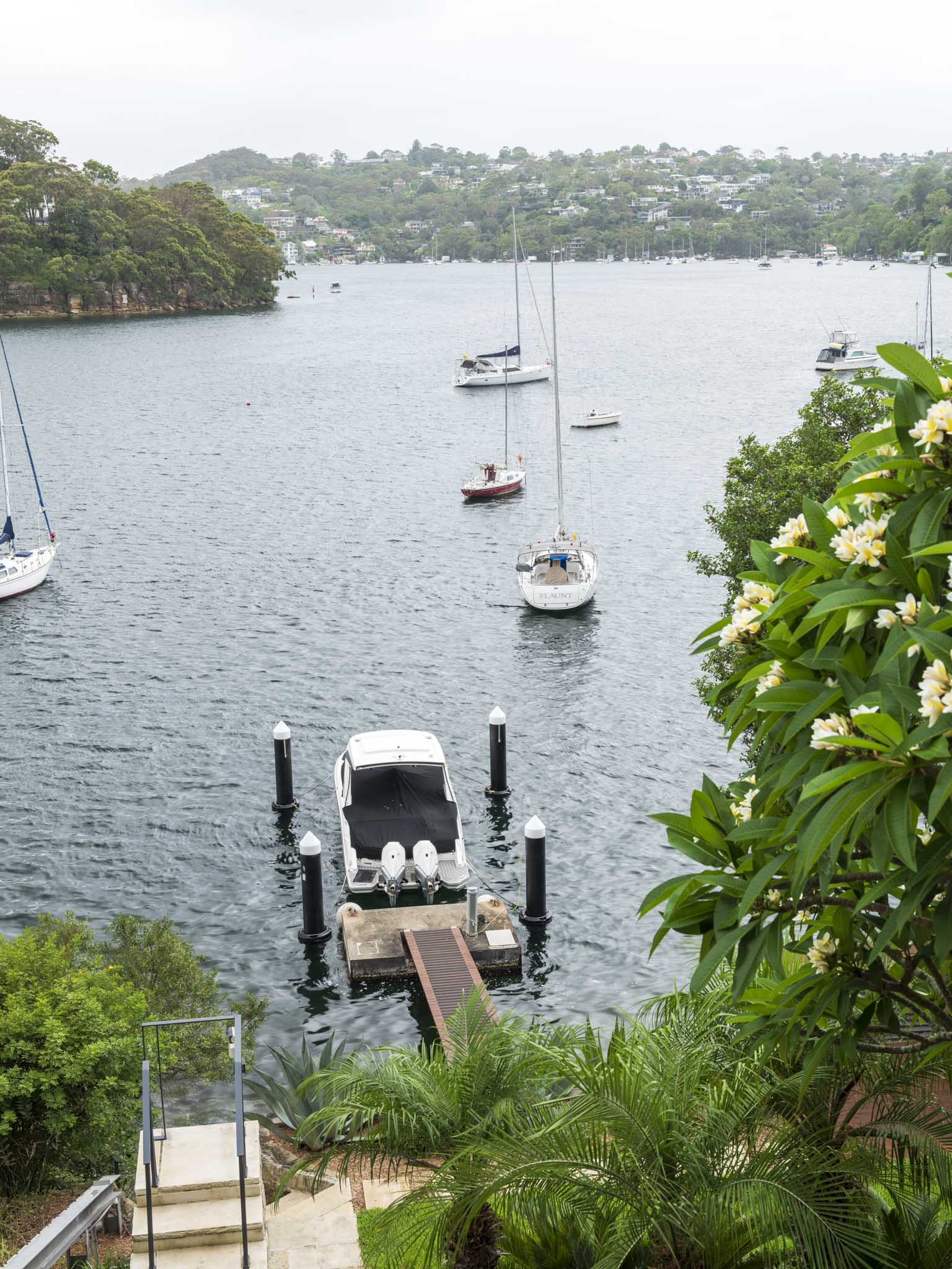 Sailors Bay by Castlepeake Architects. Photography by Tom Ferguson. View over harbour and opposite hills, covered with homes. Boats moored in water and green plants surrounding path. 