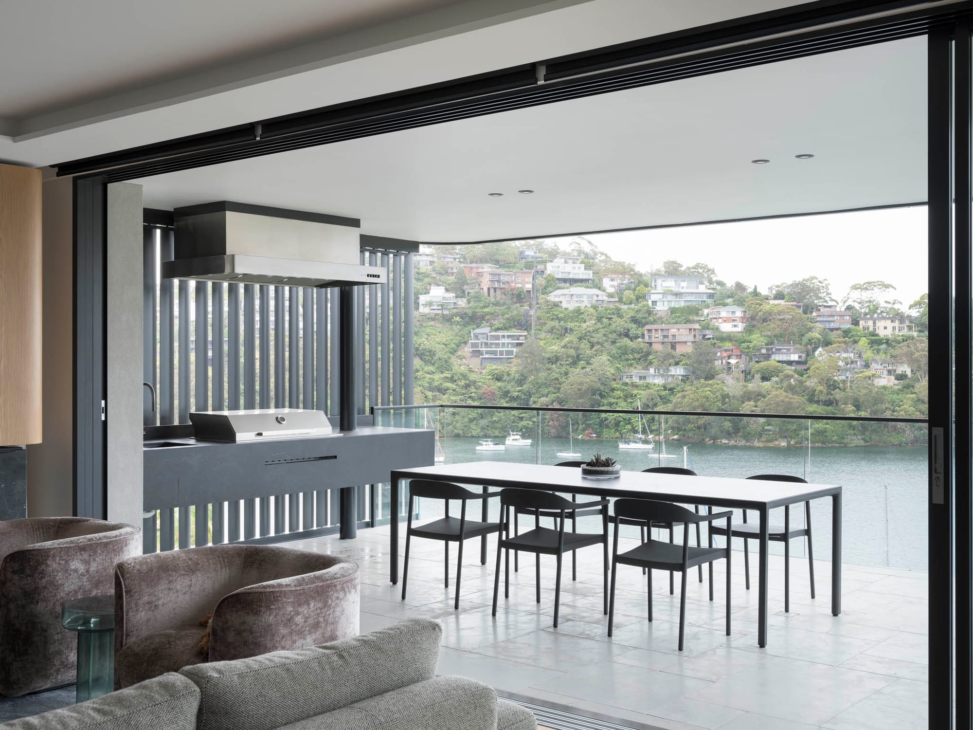 Sailors Bay by Castlepeake Architects. Photography by Tom Ferguson. Exterior dining space with glass fencing and views over water and hills, as well as outdoor barbeque to the left. 