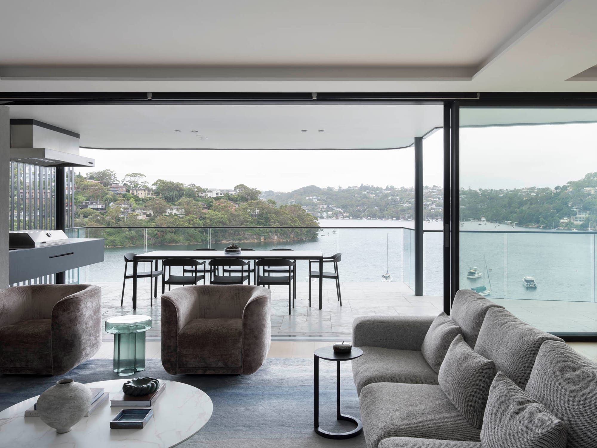 Sailors Bay by Castlepeake Architects. Photography by Tom Ferguson. Views over the harbour from open plan living space and adjoining paved balcony with glass fencing and grey couches.