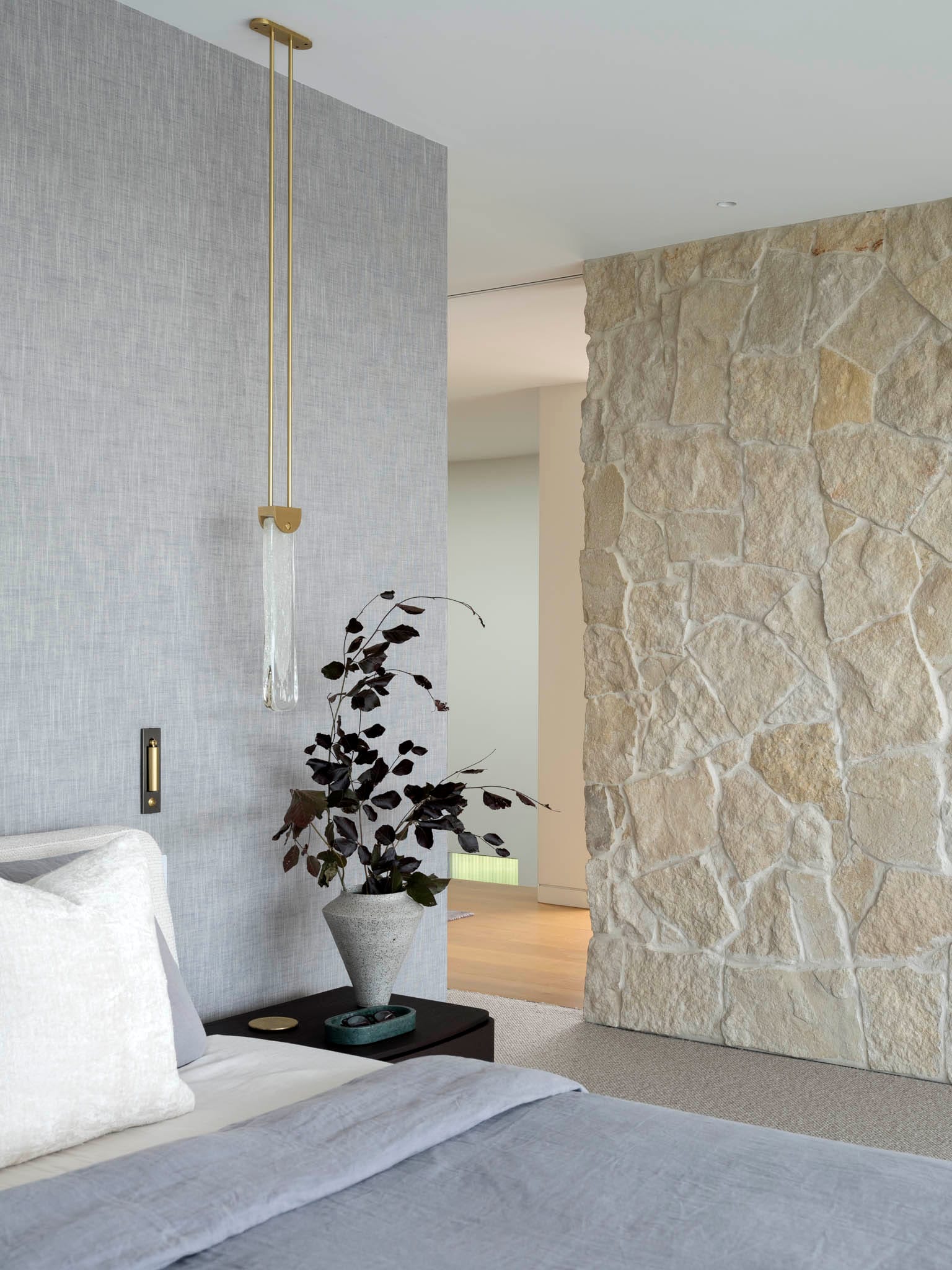 Sailors Bay by Castlepeake Architects. Photography by Tom Ferguson. Bedroom with sandstone feature wall to the right and grey linen wall to the left, with beige carpet and hanging pendant light at bedside. 