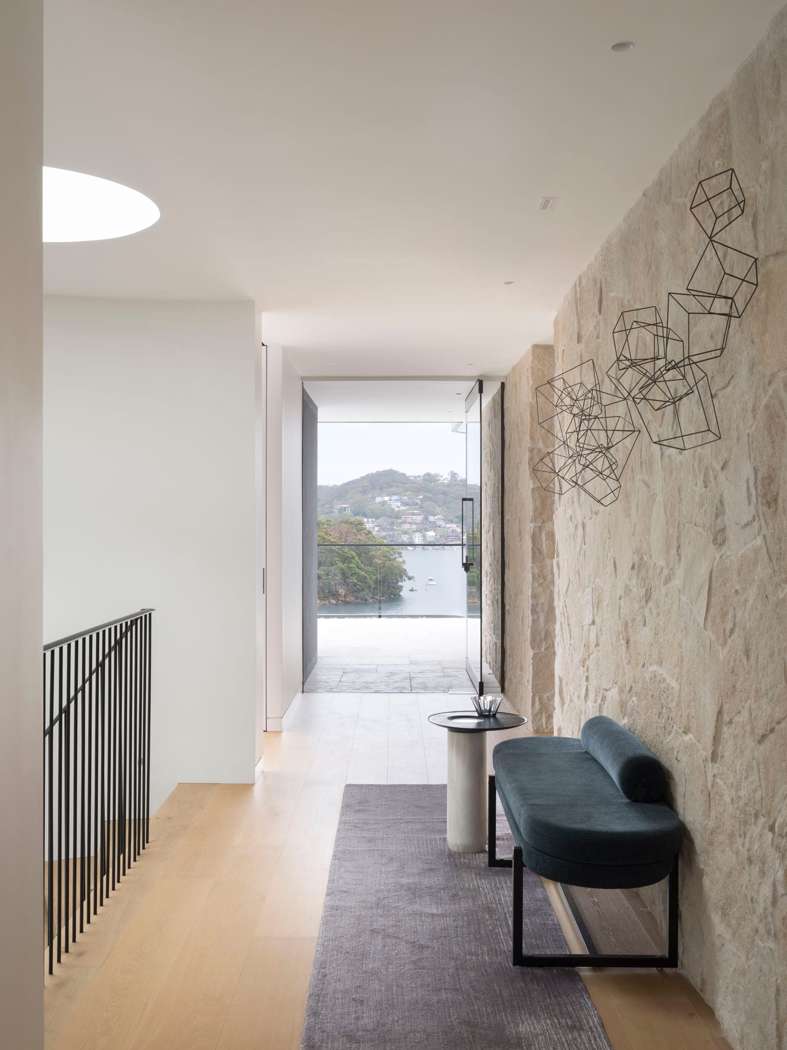 Sailors Bay by Castlepeake Architects. Photography by Tom Ferguson. Hallway leading to tiled balcony with views over water and hills, with sandstone feature wall and black metal railing around stairs. 