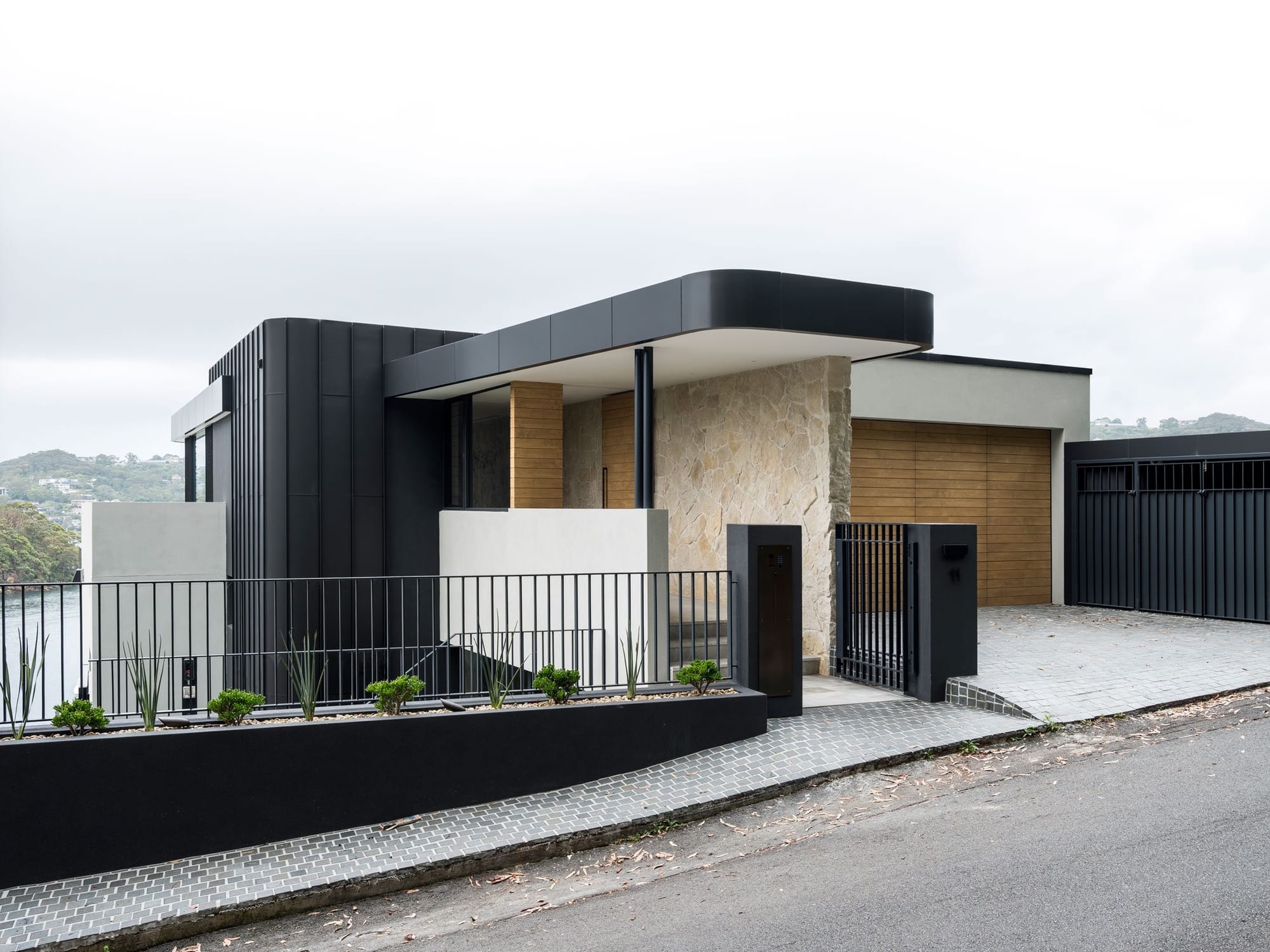 Sailors Bay by Castlepeake Architects. Photography by Tom Ferguson. Exterior front facade of home with black steel cladding, sandstone, timber and paved driveway. 