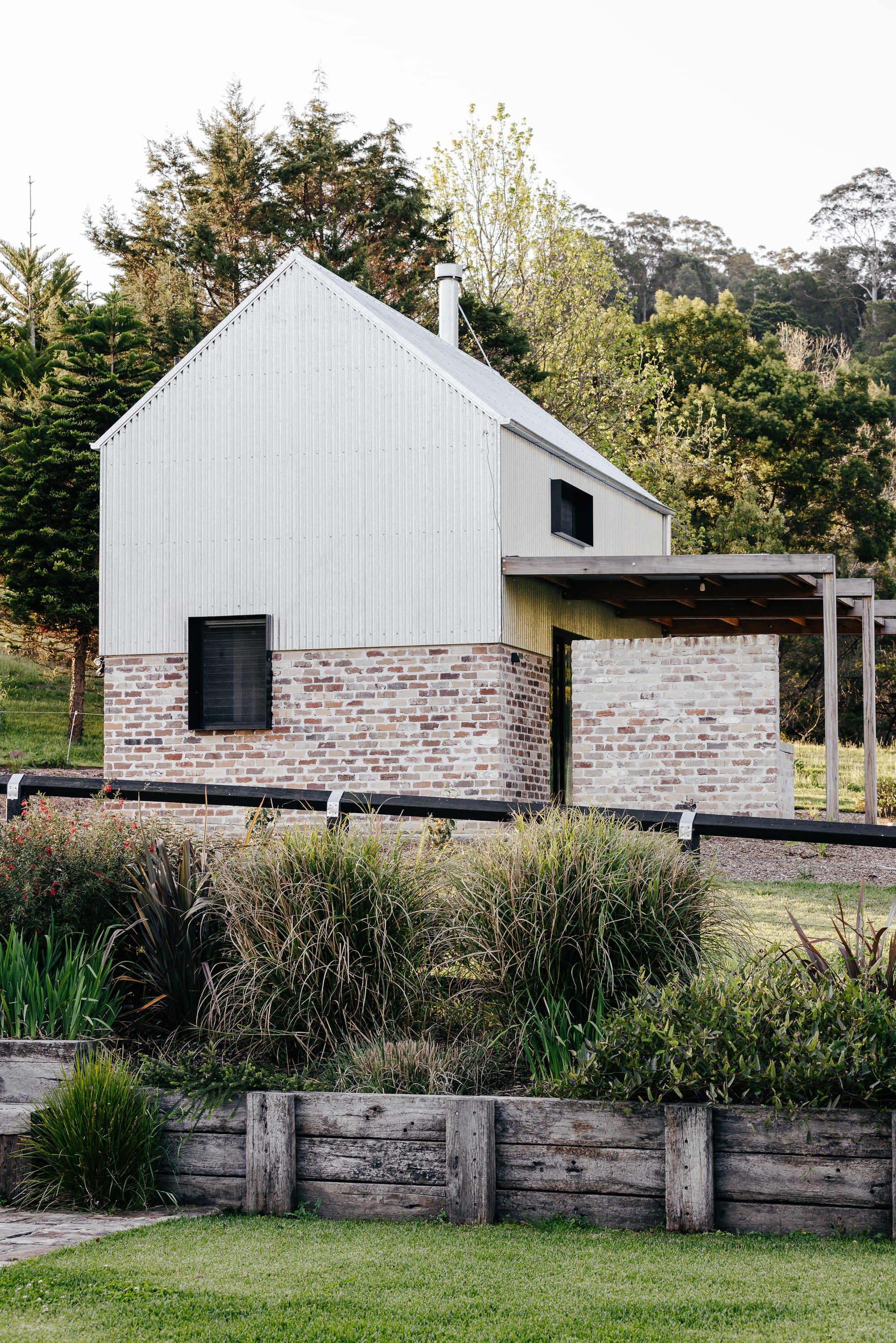 Reclaimed timber garden beds with various bushes and plants, in front of a double storey brick and tin home with simple pitched roof and timber verandah.