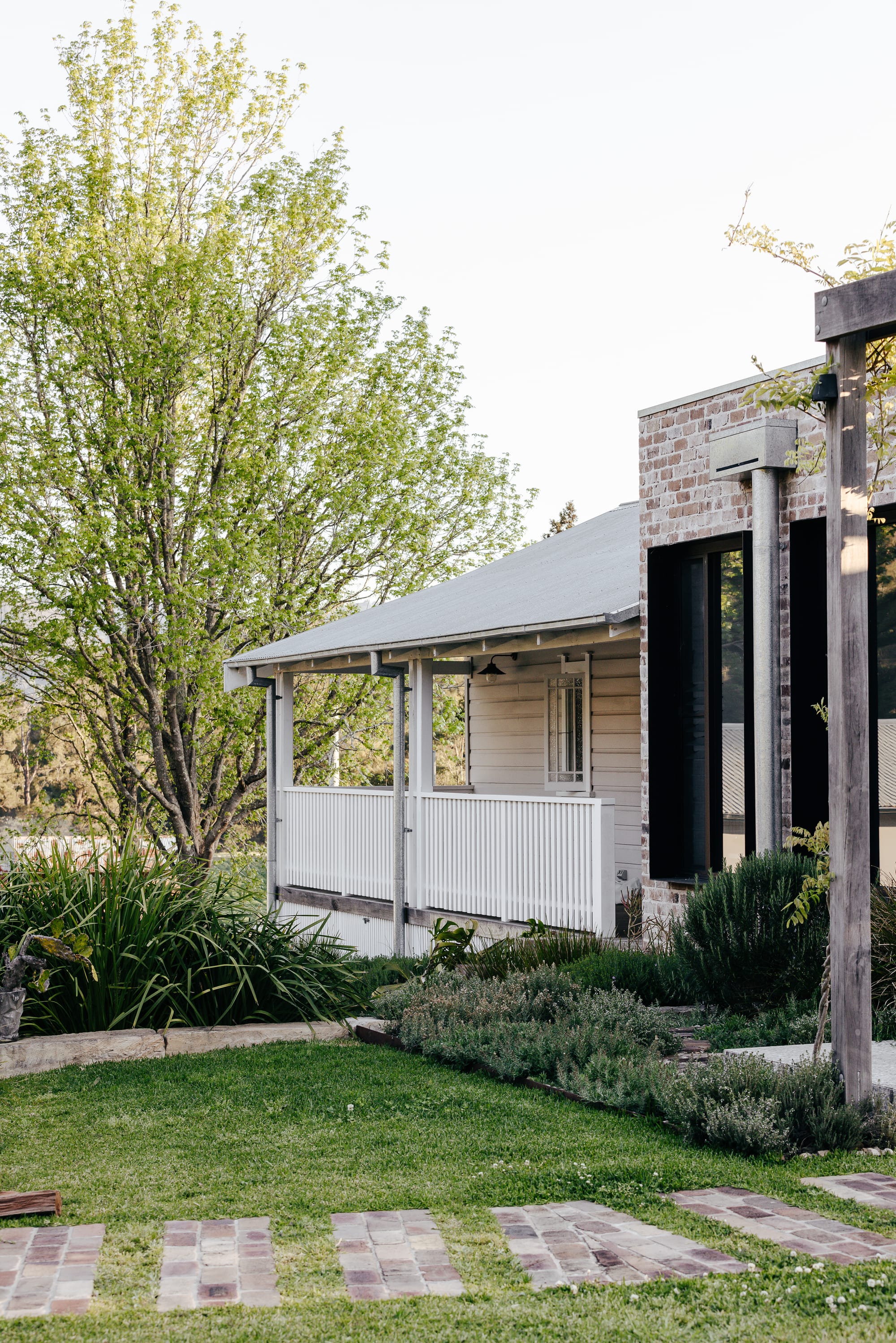 Side of home with undervocer verandah in front of timber clad facade. Brick pavers carving footpath through green grass. 