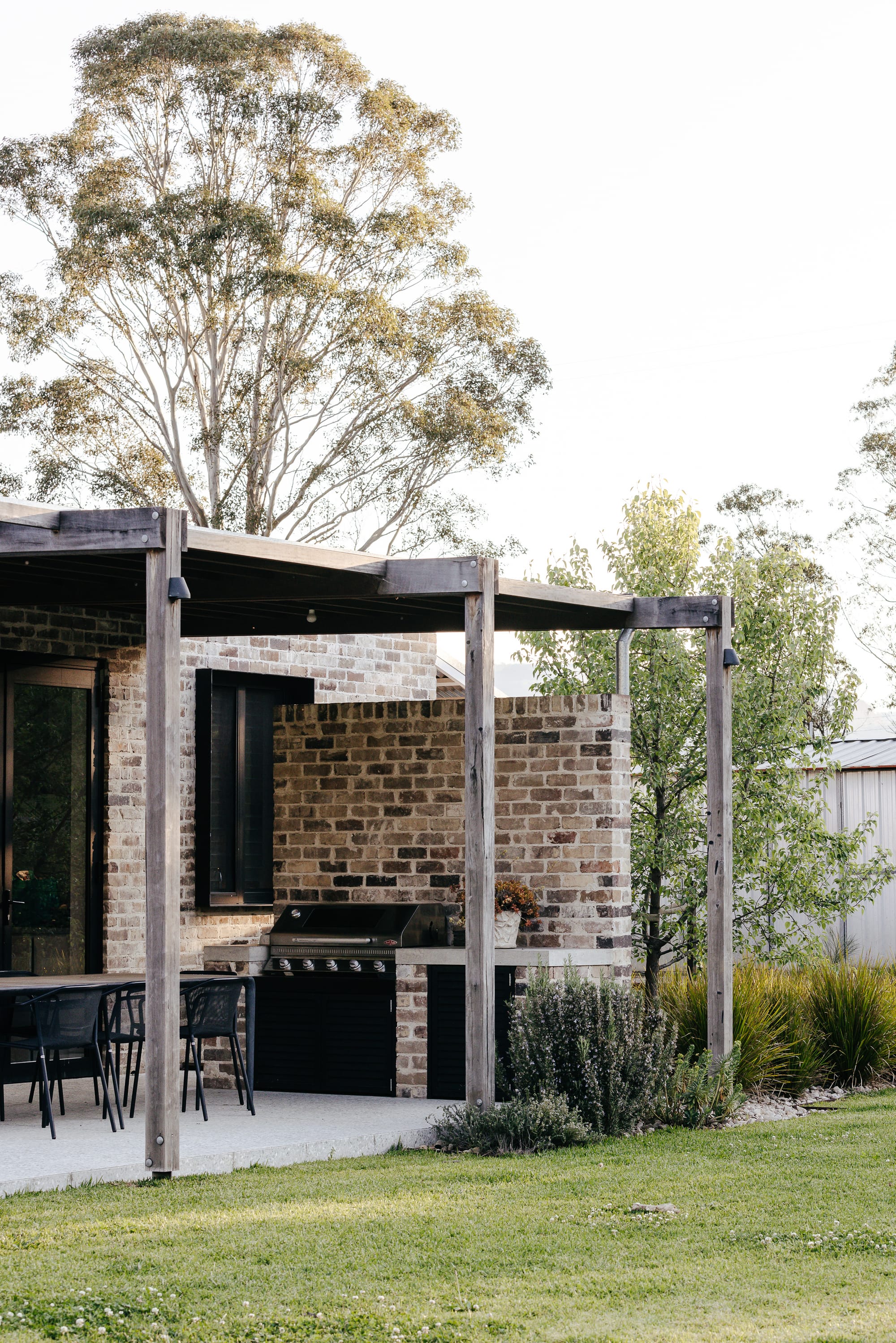 Enclosed outdoor entertaining area with brick wall, black integrated barbeque and black dining setting. Grass runs flush to concrete floor.