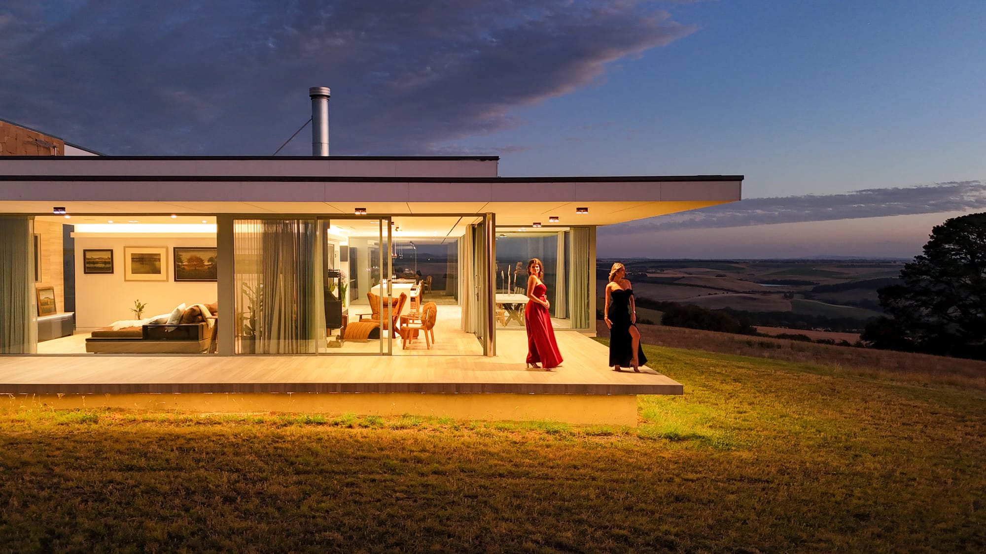 The Cloud at Sen Vineyard. Photography by Ben Little. Contemporary home at night, with warm light visible through the homes glass walls and windows. Two women in dresses standing in outdoor verandah overlooking surrounding paddocks. 