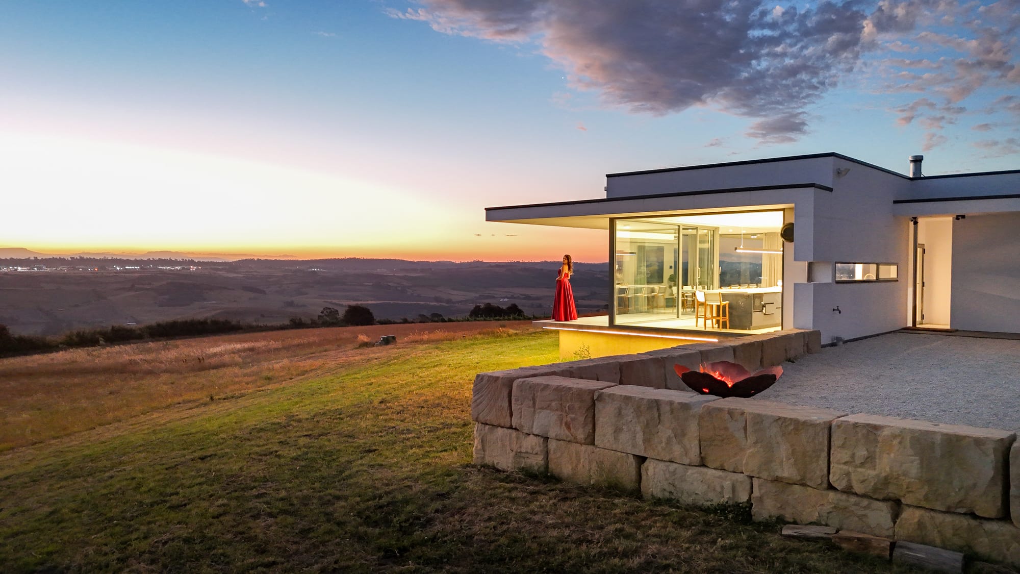 The Cloud at Sen Vineyard. Photography by Ben Little. Enclosed courtyard area with white gravel and firepit to left of contemporary white home at night, with warm light visible through full length glass walls. 