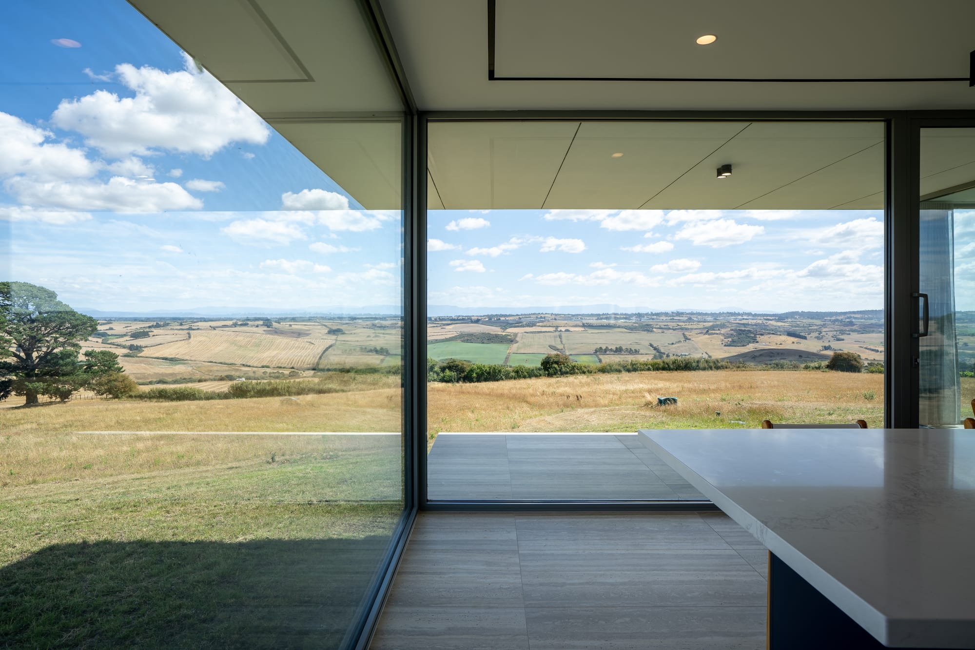 The Cloud at Sen Vineyard. Photography by Ben Little. Floor-to-ciling glass windows and doors on two corner walls of a space, overlooking rolling green paddocks. 