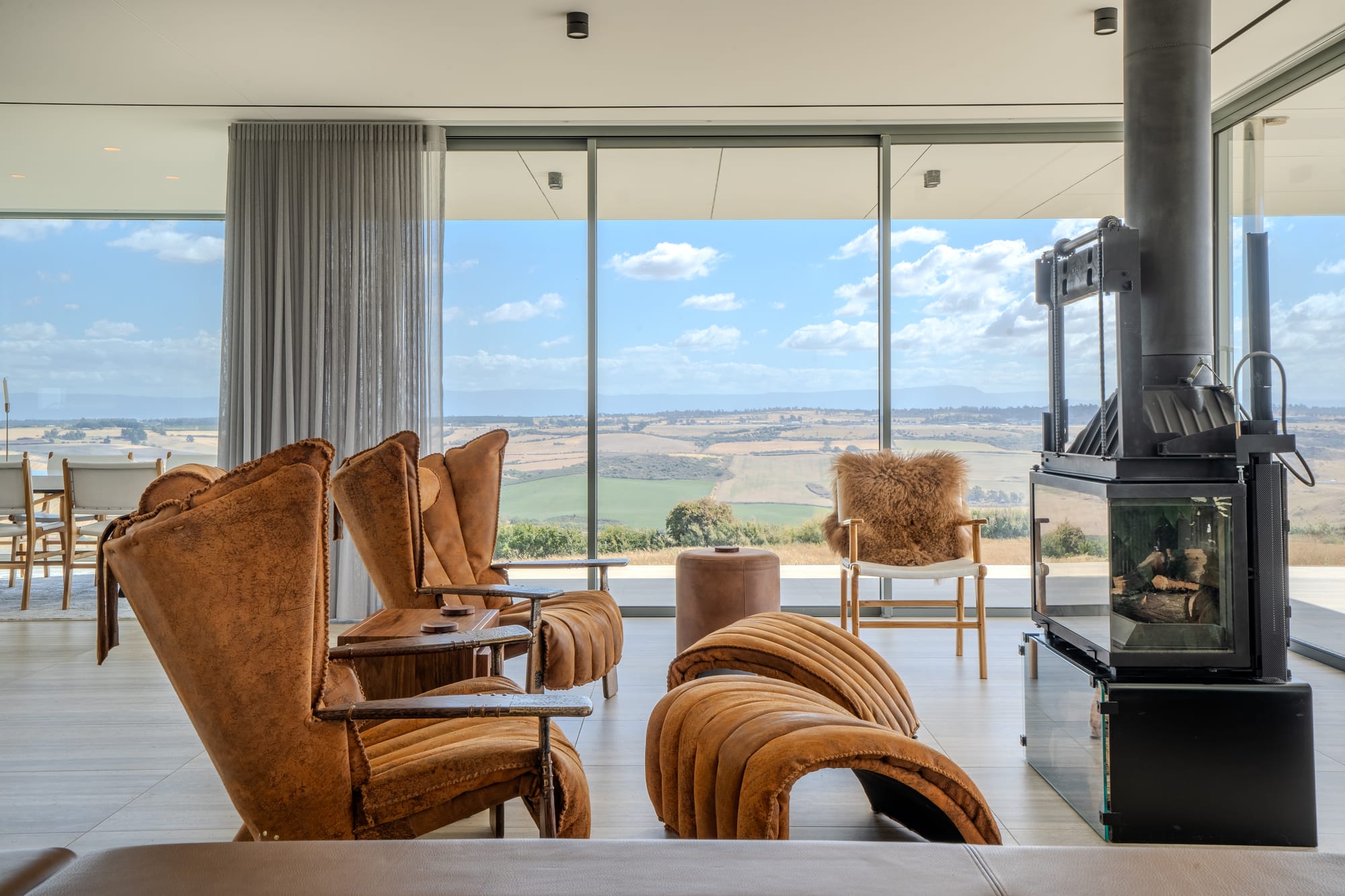 The Cloud at Sen Vineyard. Photography by Ben Little. Living space with art nouveau tan leather chairs, and a freestanding black and glass fireplace. Full length glass windows around perimeter of room overlooking paddocks. 