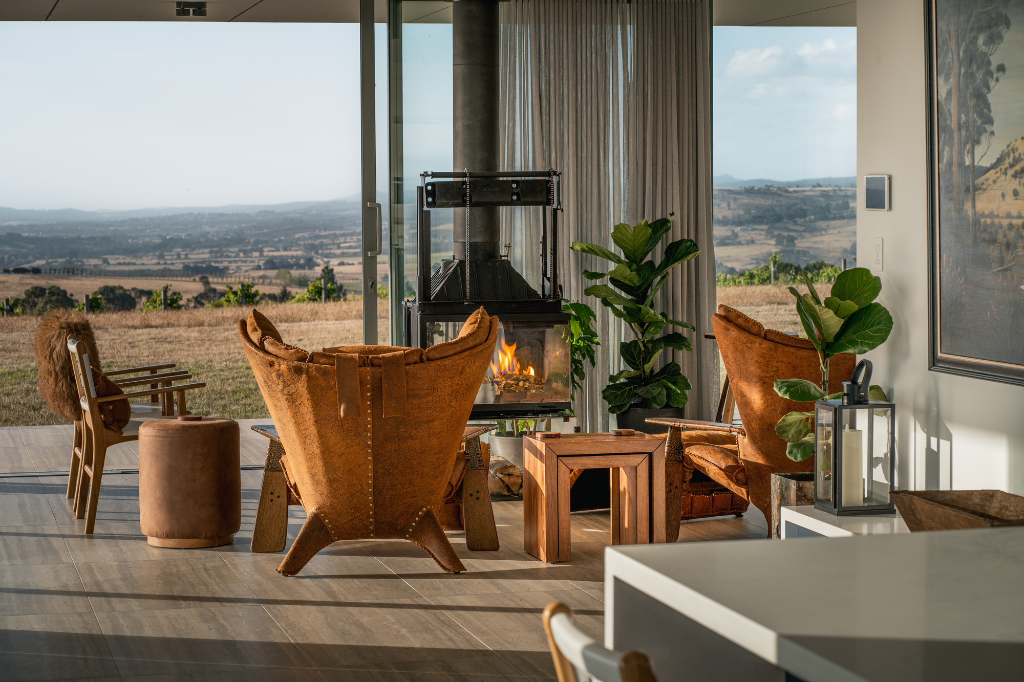 The Cloud at Sen Vineyard. Photography by Ben Little. Living space withtan leather arm chairs and timber seating, with an exposed glass fireplace and full-length windows overlooking rolling hills. 