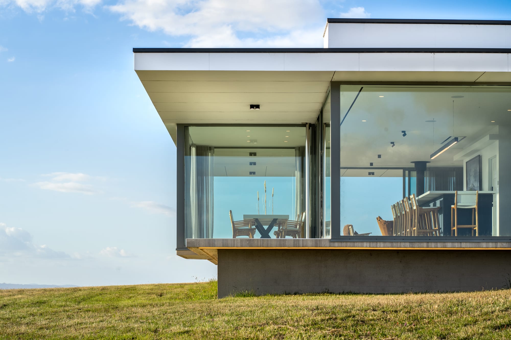 The Cloud at Sen Vineyard. Photography by Ben Little. Low angle of contemporary home on elevated foundation, covered entirely in full length glass windows and doors. Kitchen and dining area visible through glass. 