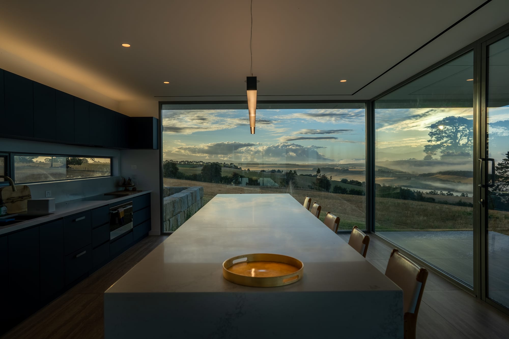 The Cloud at Sen Vineyard. Photography by Ben Little. Modern kitchen with ling white island bench and stool in front of full-length glass window with scenic views. Black cabinetry and pendant light. 