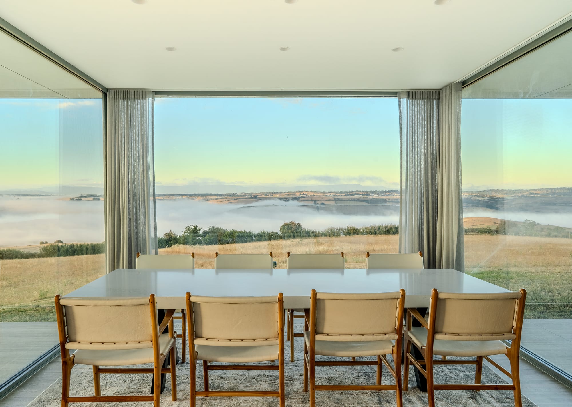 The Cloud at Sen Vineyard. Photography by Ben Little. Dining area with full length glass windows on 3 walls, overlooking clouds and paddocks. White dining table with white leather and timber chairs. 