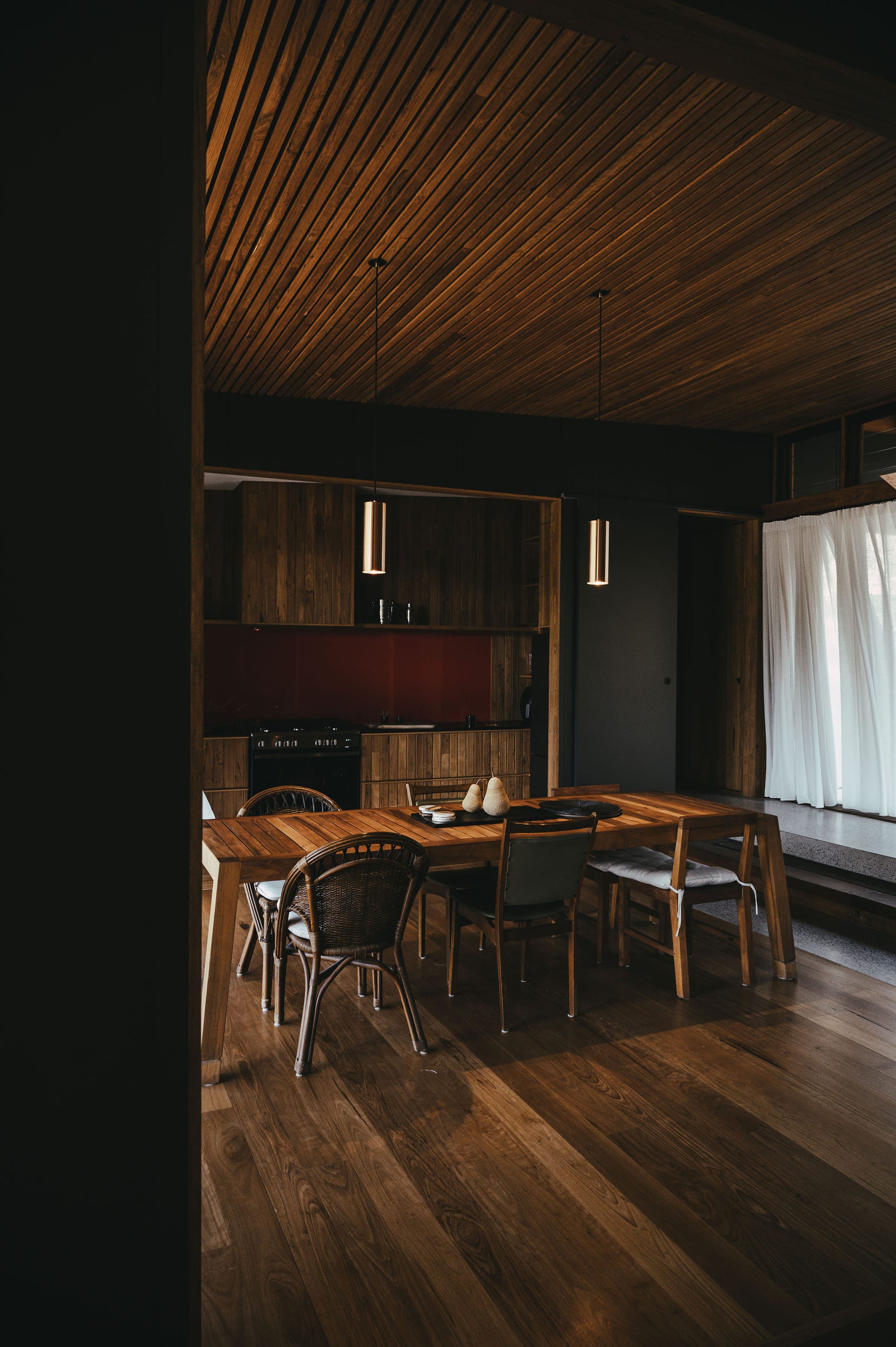 The Summer House at Little Valley Farm. Photography by Milou Hofman Photography. Open plan dining and kitchen space with timber floors and joinery, red splashback and mismatched dining chairs.