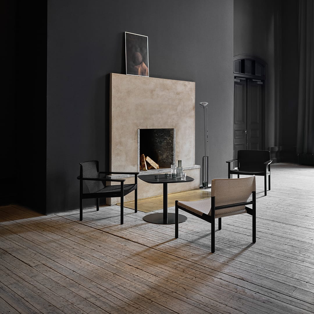 Evo Chair by EFG. Dark, moody room with black walls and grey stone fireplace, with three timber and knit chairs arranged around a black side table. 