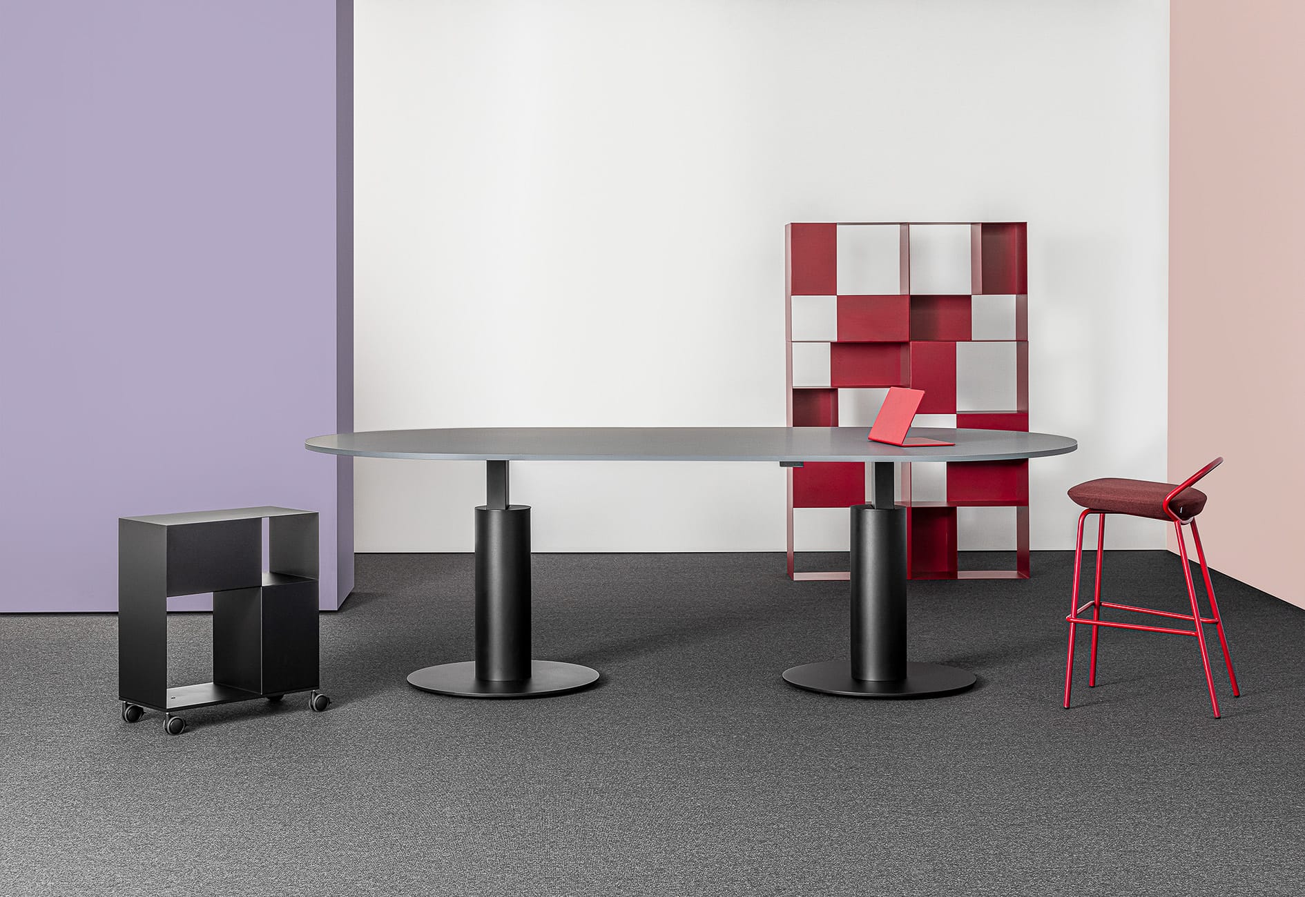 Follow Meeting Table by Mara SRL.  Black work desk with rounded legs and edges in office space with local and white walls, grey carpets, and red chairs and storage units.