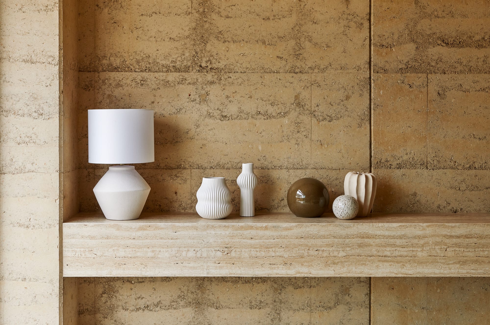 Homewares by GlobeWest from Collection 2025. Travertine floating bench with various homewares on top and rammed earth walls behind. 