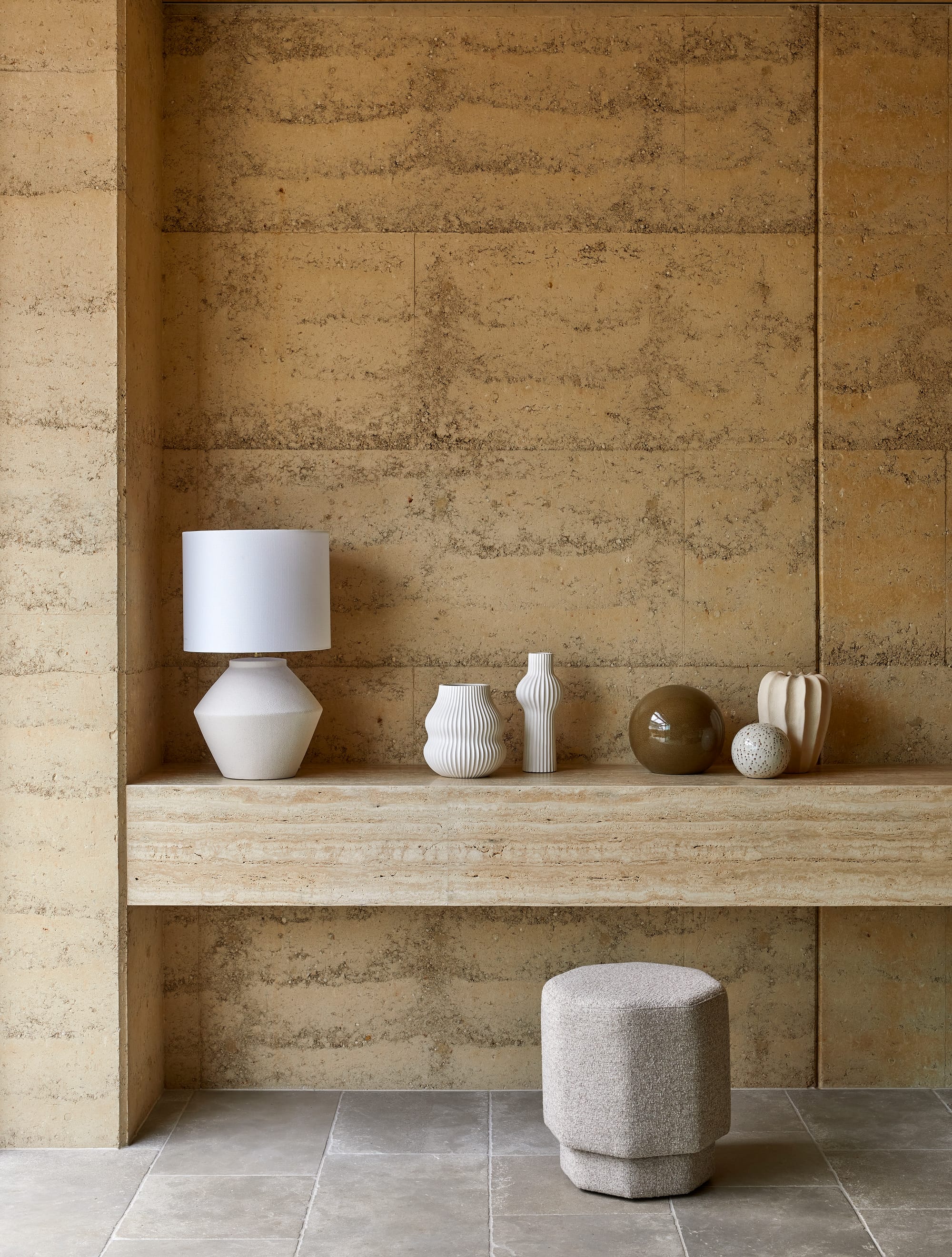 Homewares by GlobeWest from Collection 2025.  Marble travertine floating display bench with multiple vases and lamps on it in front of rammed earth wall.